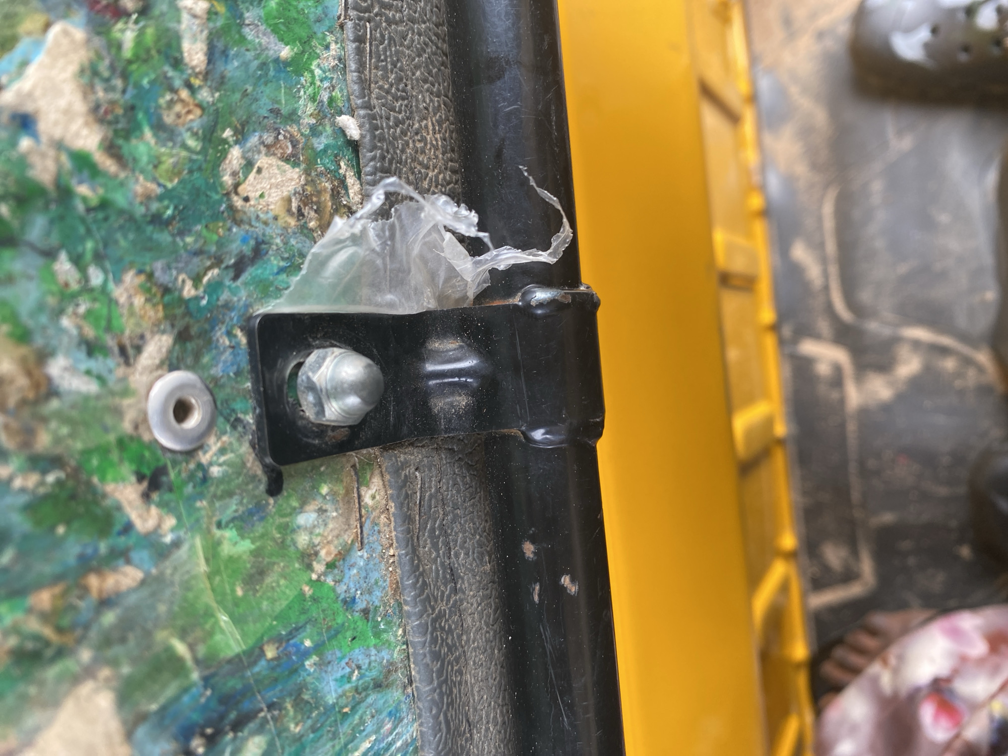 The image shows a close-up of a black metal bar attached to a textured, multicolored surface that appears to be a composite material, possibly wood or plastic, with paint or pigment embedded in it. A silver bolt and nut secure the metal bar to the surface. A piece of clear plastic sheeting is caught under the bolt. To the right of the metal bar is a bright yellow object, likely another part of the structure or vehicle. The background is blurred, but suggests an outdoor or workshop setting with a dark, textured surface and a hint of a patterned fabric. The scene is brightly lit, suggesting daylight. There are no visible people or text. The overall impression is one of wear and tear, with the multicolored surface showing signs of age and use, and the plastic sheeting indicating a makeshift repair or covering. The context of Jalingo, Nigeria, might suggest this is part of a vehicle, a piece of equipment, or a structure found in that region.