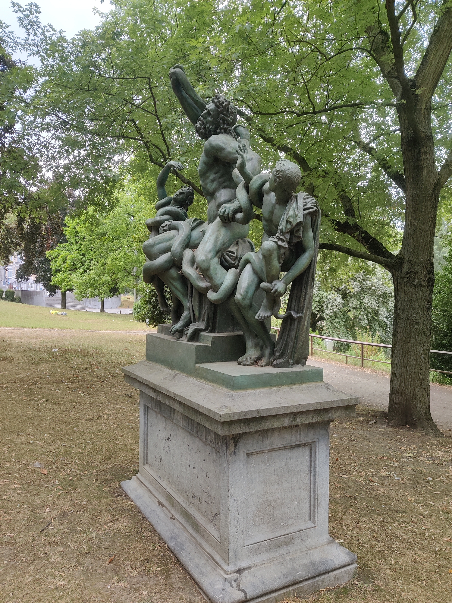 The image depicts a bronze statue on a stone pedestal situated in a park. The statue portrays a mythological scene with several figures and serpents, rendered in a dynamic and dramatic style. The primary figures appear to be a muscular male figure being attacked by snakes, with other figures interacting with the scene. The bronze has a green patina, indicating age and exposure to the elements. The stone pedestal is rectangular with decorative moldings.
The park setting is characterized by a grassy area with scattered dry leaves, a dirt path, and lush green trees in the background. In the distance, some buildings are visible on a slope, suggesting an urban environment.
This statue is known as "Laocoön and His Sons" and is located in the Parc de la Citadelle in Liège, Belgium. The statue is a replica of the Hellenistic sculpture discovered in Rome in 1506. The park itself offers panoramic views of the city and the Meuse river valley.