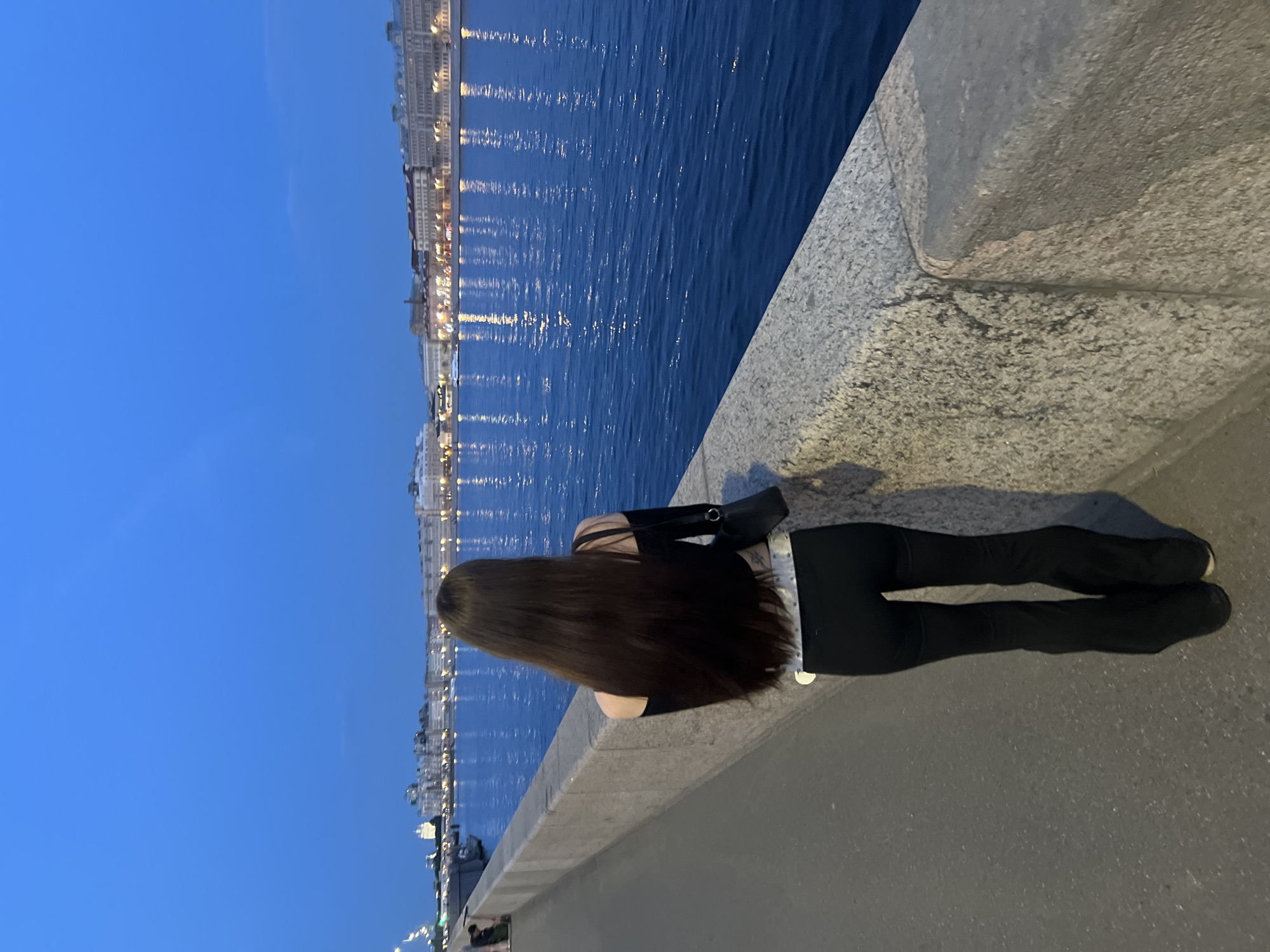 A rear view of a young woman with long, dark hair stands on a paved walkway next to a body of water at dusk. She is wearing an off-the-shoulder black top and black flared pants. A black purse is slung over her right shoulder. The walkway is bordered by a stone embankment. Across the water, a city skyline is visible with buildings illuminated by lights. The sky is a deep blue, indicating it is twilight.