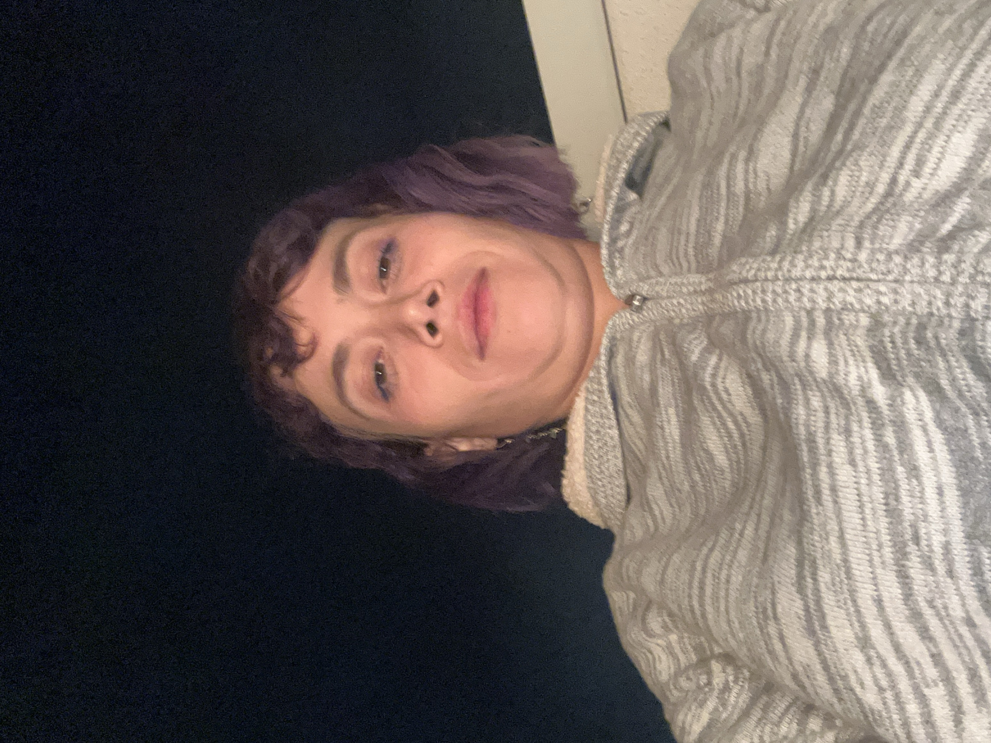 A low-angle, head-and-shoulders portrait captures an adult female with light skin and wavy, shoulder-length purple hair, including bangs. She looks directly at the camera with a neutral expression, displaying slight eye makeup and a visible double chin. She is wearing a grey and white patterned knitted jacket with a central zipper and a cream-colored sherpa-lined collar. A silver-colored earring is visible on her left ear. The background is predominantly dark, indicating a nighttime setting, with a light-colored, possibly architectural, horizontal element visible in the lower right section behind her right shoulder. The scene is located in the city of Los Angeles, United States of America.