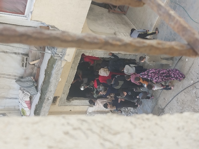 An overhead view captures a crowd, predominantly children and young adults, gathered around the damaged ground-level entrance of a building in Khan Yunis, Palestinian Territory. The building structure shows signs of damage, with a large, irregular opening serving as the entrance. Above this entrance, a partially collapsed concrete ledge supports a rolled mattress and several cinder blocks. Inside the opening, an individual holds a large metal pot or bowl, seemingly distributing contents to people clustered around them, many of whom are holding small white buckets or containers. Outside the entrance, on uneven, debris-strewn ground, an adult woman in a dark patterned dress stands with several small children. Other individuals are positioned around the entrance. A rusted metal bar in the foreground partially obscures the right side of the frame.