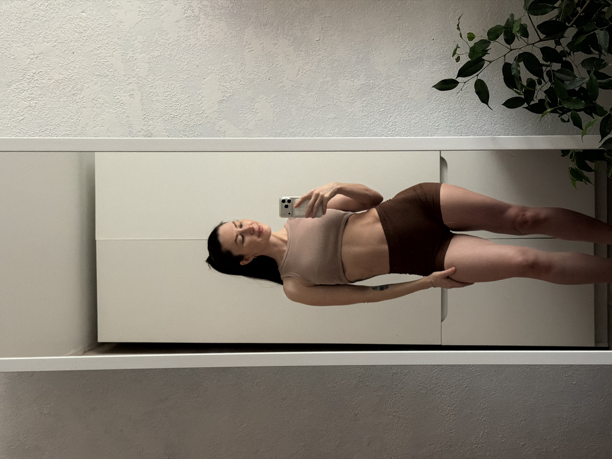 A woman in a beige top and brown shorts is seen in a mirror. She is holding a smartphone in her right hand and looking at the screen. The mirror reflects a white wall and part of a green plant. The background includes a textured white wall and a white storage unit with vertical panels. The location is an interior space, potentially in a home or apartment. There is no information indicating the location to be Perm, Russia.
