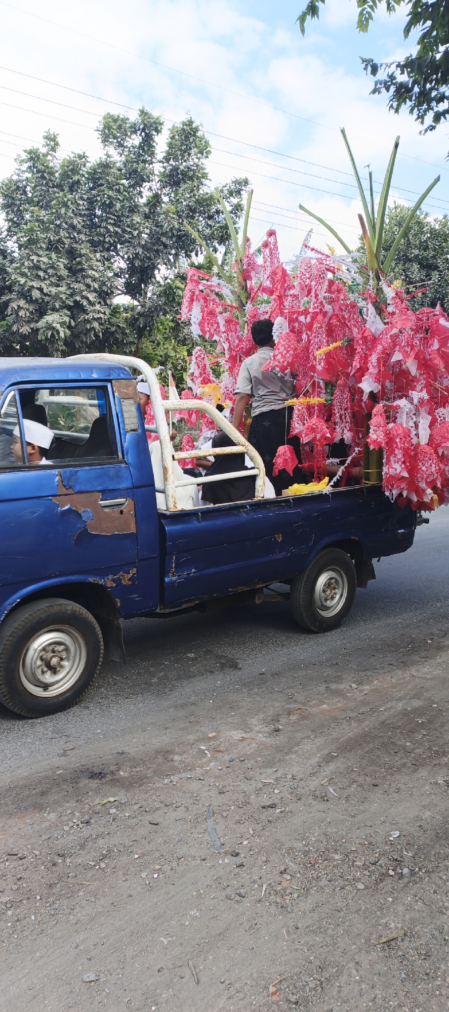 A weathered blue pickup truck, with significant rust on its side panel, is parked on a gravelly, dusty road. The truck bed is filled with people and what appears to be decorative elements, primarily red and white paper ornaments hanging from tall stalks. In the foreground, the driver's side front tire and wheel are clearly visible. In the background, lush green trees line the road, with a cloudy sky overhead. There are power lines visible in the distance. The scene suggests a procession or cultural event.