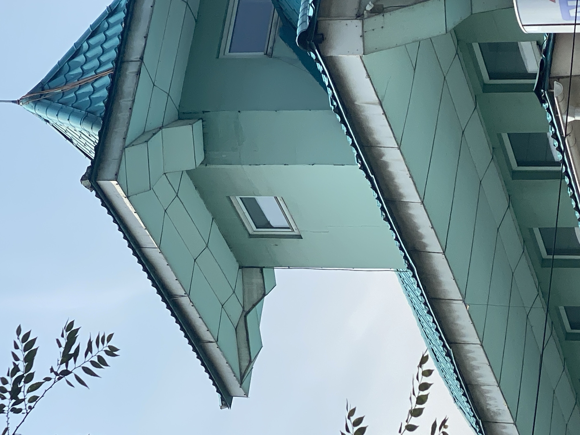 The image shows the upper portion of a building with a teal-colored tiled roof and light green walls. The building has several windows, some of which are visible. The architecture appears to be modern, with sharp angles and geometric shapes in the roof structure and eaves. There are also tree branches with dark leaves in the upper left and lower left portions of the frame, silhouetted against a light blue sky. No people are visible in the image. The scene is an outdoor view of a building's exterior.