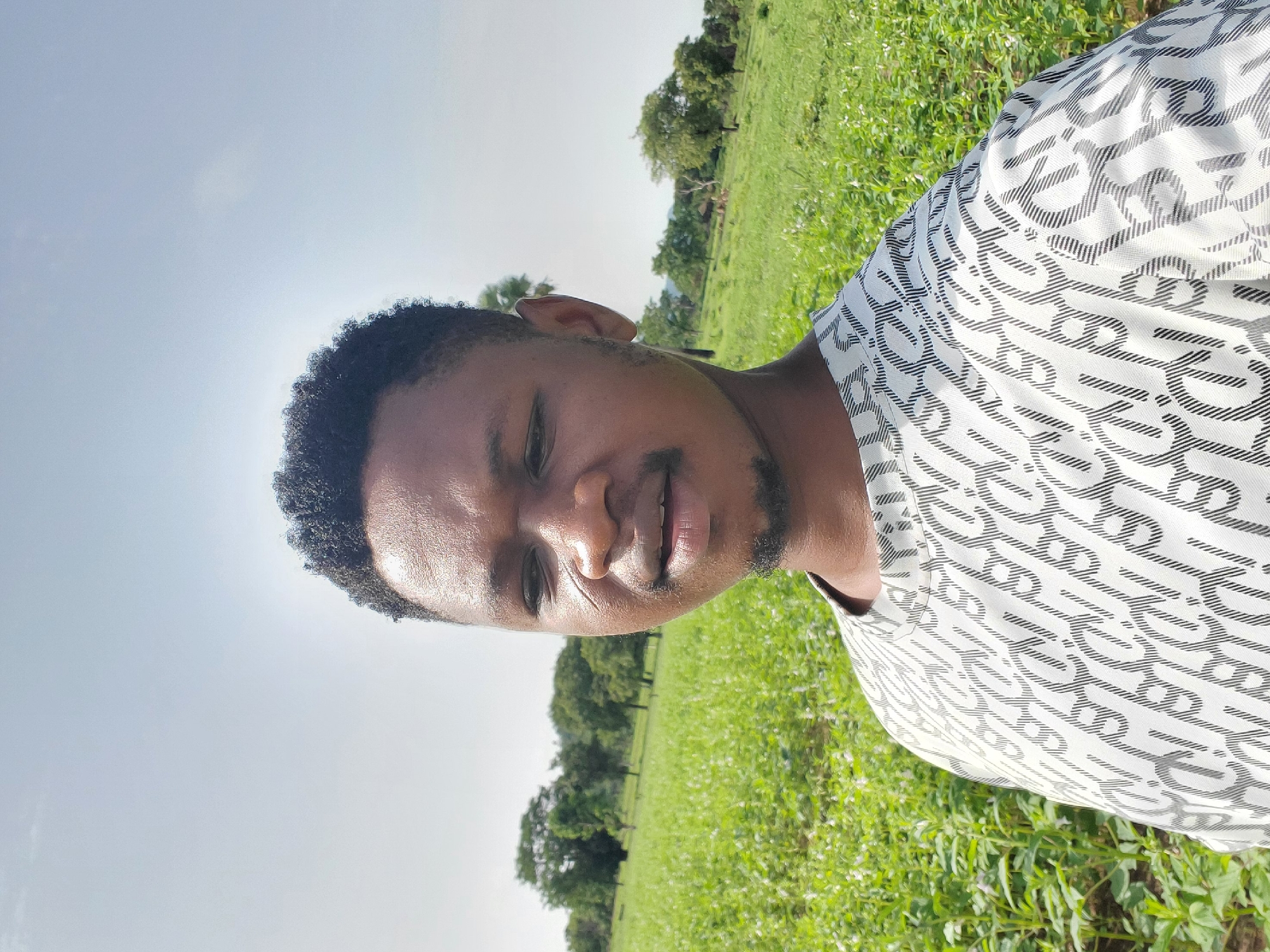 A medium shot captures a man against the backdrop of a grassy field beneath a clear sky. The man has dark hair and a goatee. He is wearing a white shirt with a black pattern. The field is filled with green vegetation, and trees are visible in the background. The location is Mararaba, Nigeria.
