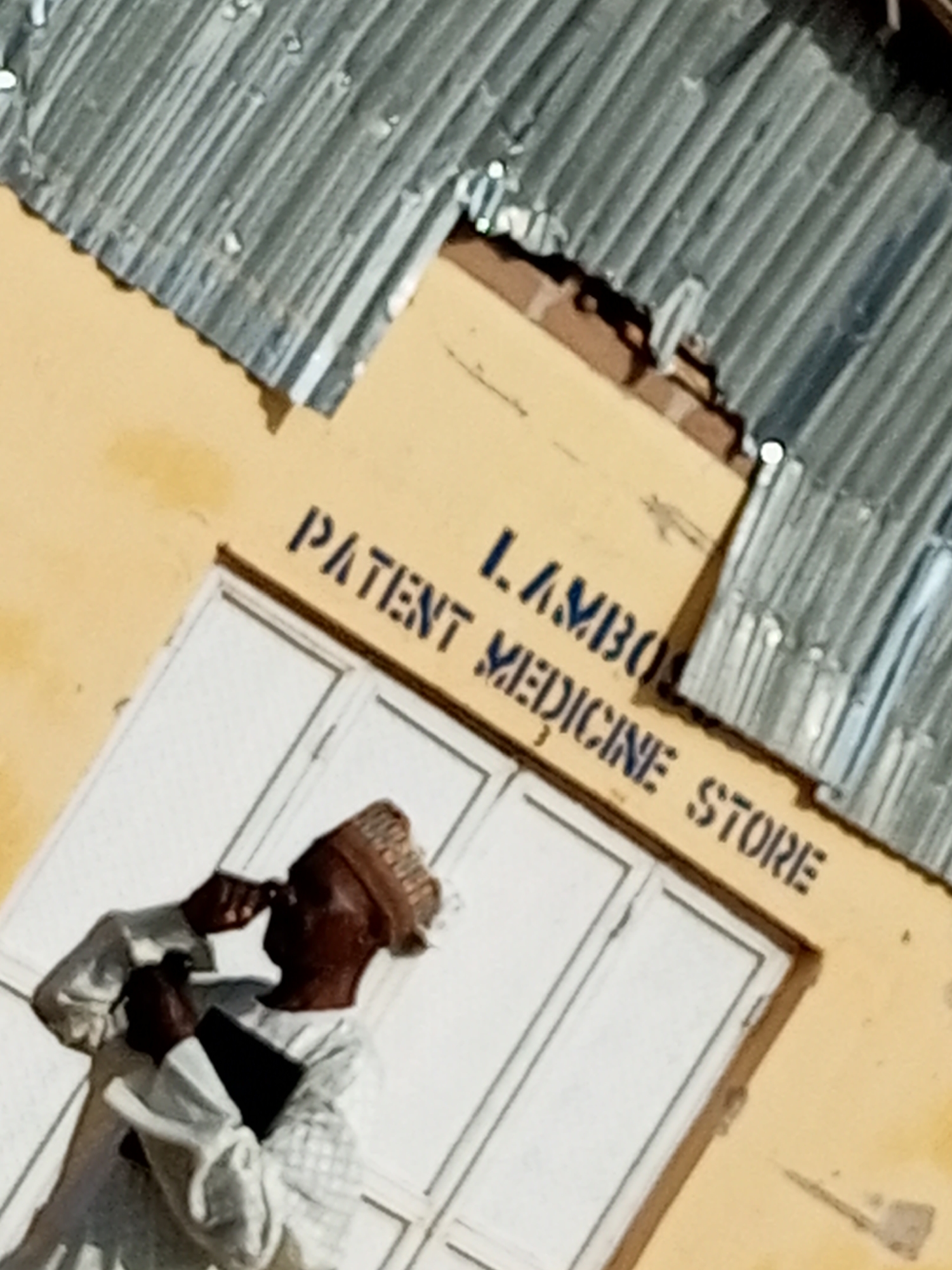 The image shows the facade of a business in Gombe, Nigeria, identified by the signage "LAMBO PATENT MEDICINE STORE" painted in dark blue letters on a light yellow wall. Above the store, a corrugated metal roof is visible, showing signs of weathering. The store entrance has double white doors. A person, appearing to be a man wearing a traditional cap and a light-colored patterned garment, stands in front of the store, holding a dark object, possibly a book or tablet, and is looking upwards and to the side, with one hand near his face. The scene appears to be outdoors, likely in a street or market area within the city of Gombe.