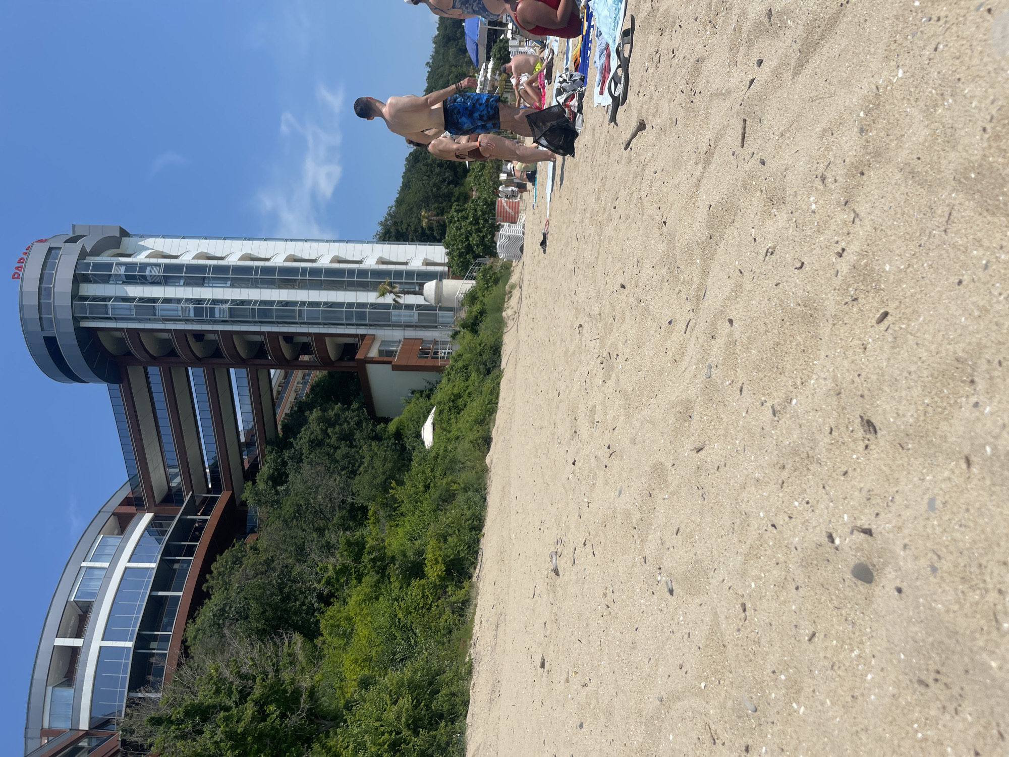 This media file captures a sunny beach scene, likely at a resort in Sveti Vlas, Bulgaria. The foreground is dominated by light-colored sand, scattered with small pebbles and shell fragments. Several people are enjoying the beach; in the midground, a man in blue patterned swim shorts stands with his back to the camera, next to another person in a brown swimsuit. Further back, other individuals are visible lounging on towels and near beach chairs, with a blue umbrella partially seen.
To the left, a modern, multi-story building, likely a hotel or resort, rises prominently. Its architecture features a distinctive cylindrical top section with large windows and a curved lower facade, made of glass, concrete, and horizontal dark panels. Lush green trees and dense vegetation separate the sandy beach from the building. The word "PARADISE" is visible in red letters on the cylindrical part of the building near the top. The sky above is clear and bright blue with a few wispy clouds, indicating good weather. The overall activity suggests people relaxing and enjoying a leisurely day at a coastal vacation spot during bright daylight hours.