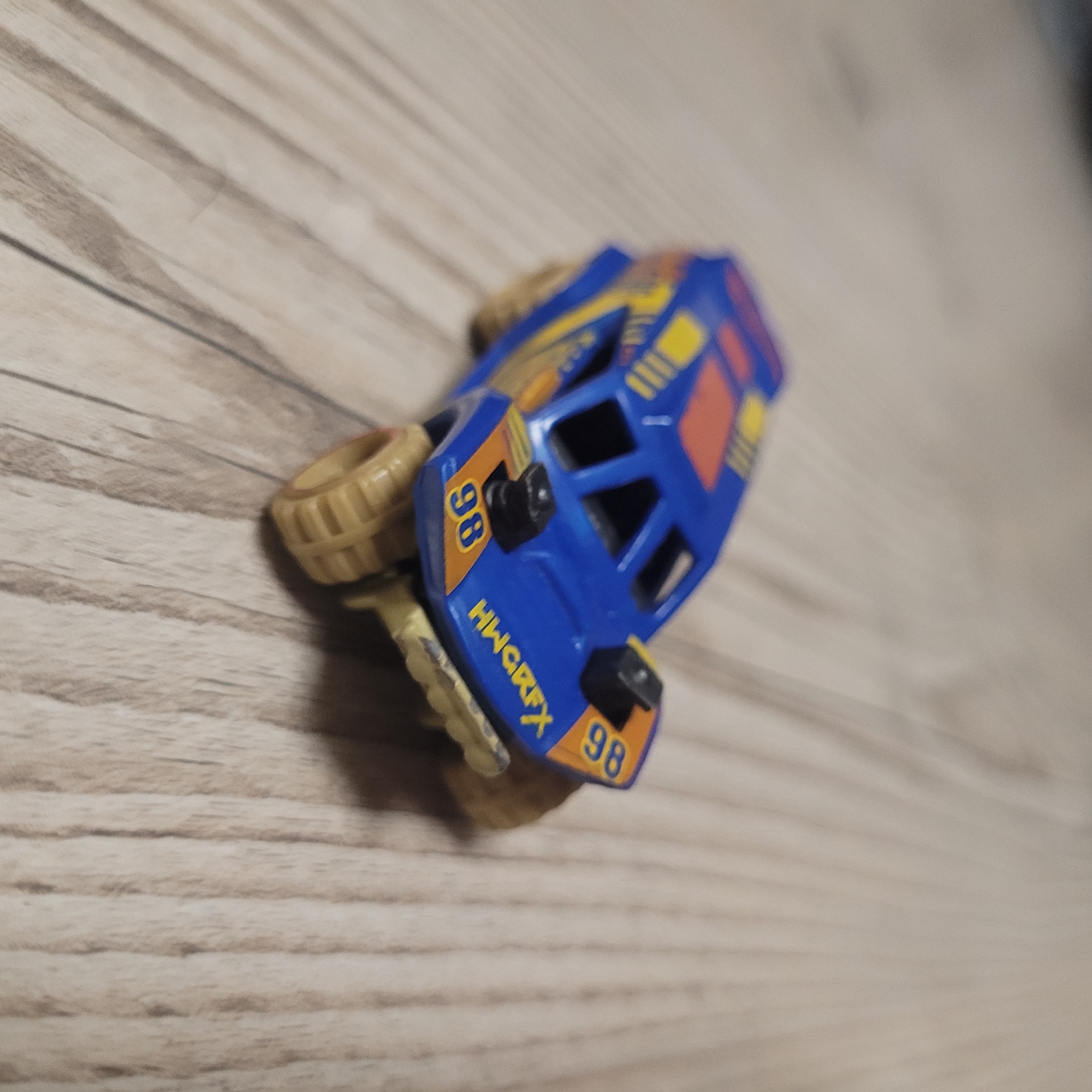 A blue and orange toy off-road vehicle is positioned on a light-colored wooden surface. The vehicle has yellow accents and the number "98" is visible on both sides of the front. The word "HWGRFX" is printed in yellow on the front of the vehicle. The vehicle has large, tan-colored wheels. The surface it rests on appears to be a floor or tabletop with a wood grain pattern. The lighting suggests it is indoors.