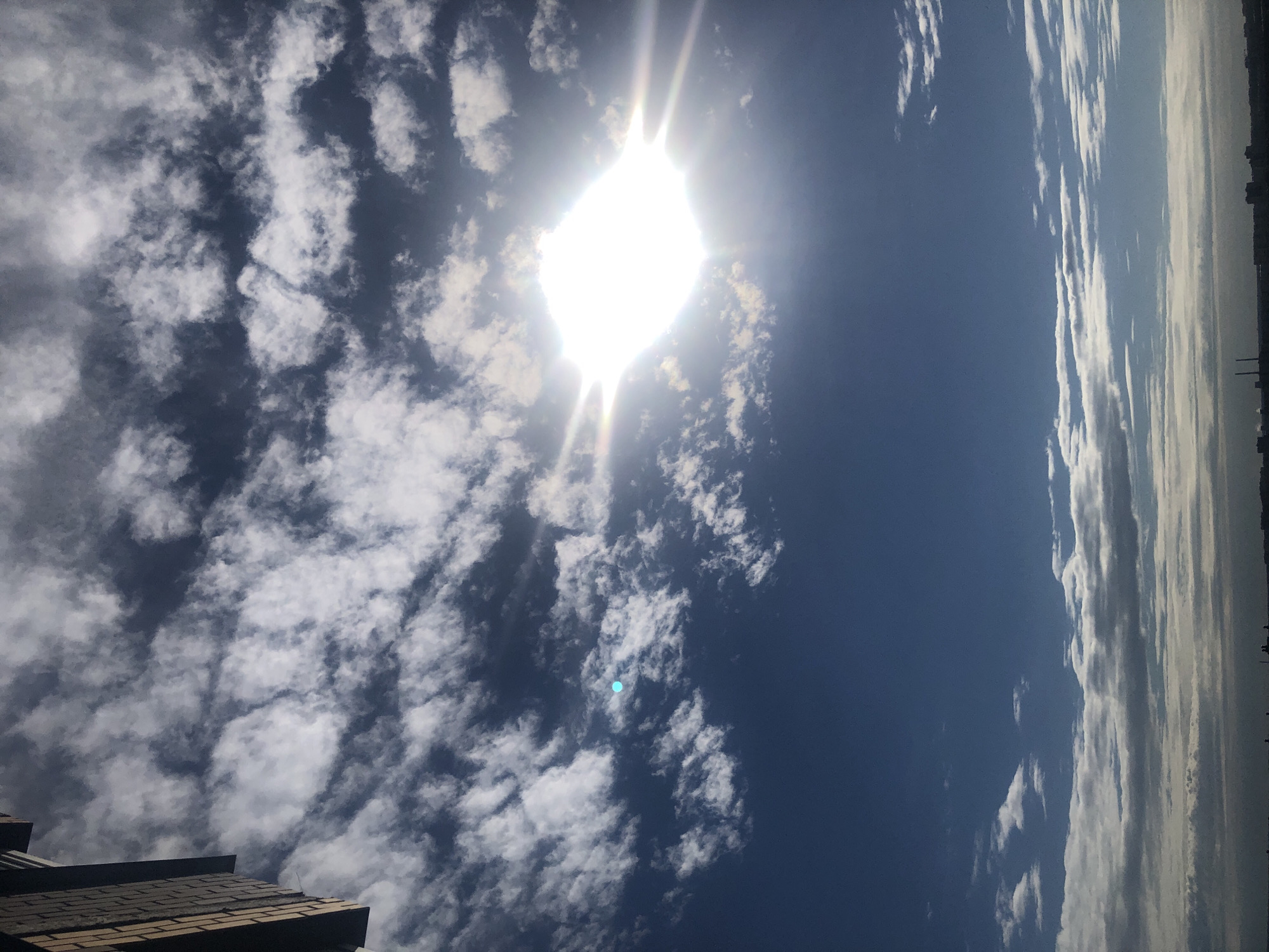 The image shows a bright sun obscured by clouds, creating a lens flare effect. The sky is a deep blue with scattered white and dark gray clouds. In the upper left corner, a portion of a brick building is visible, suggesting an urban environment. The horizon in the distance shows silhouettes of buildings and possibly industrial structures. The scene appears to be captured from a high vantage point, overlooking a cityscape. The overall lighting suggests it is daytime, with the sun being the dominant light source.