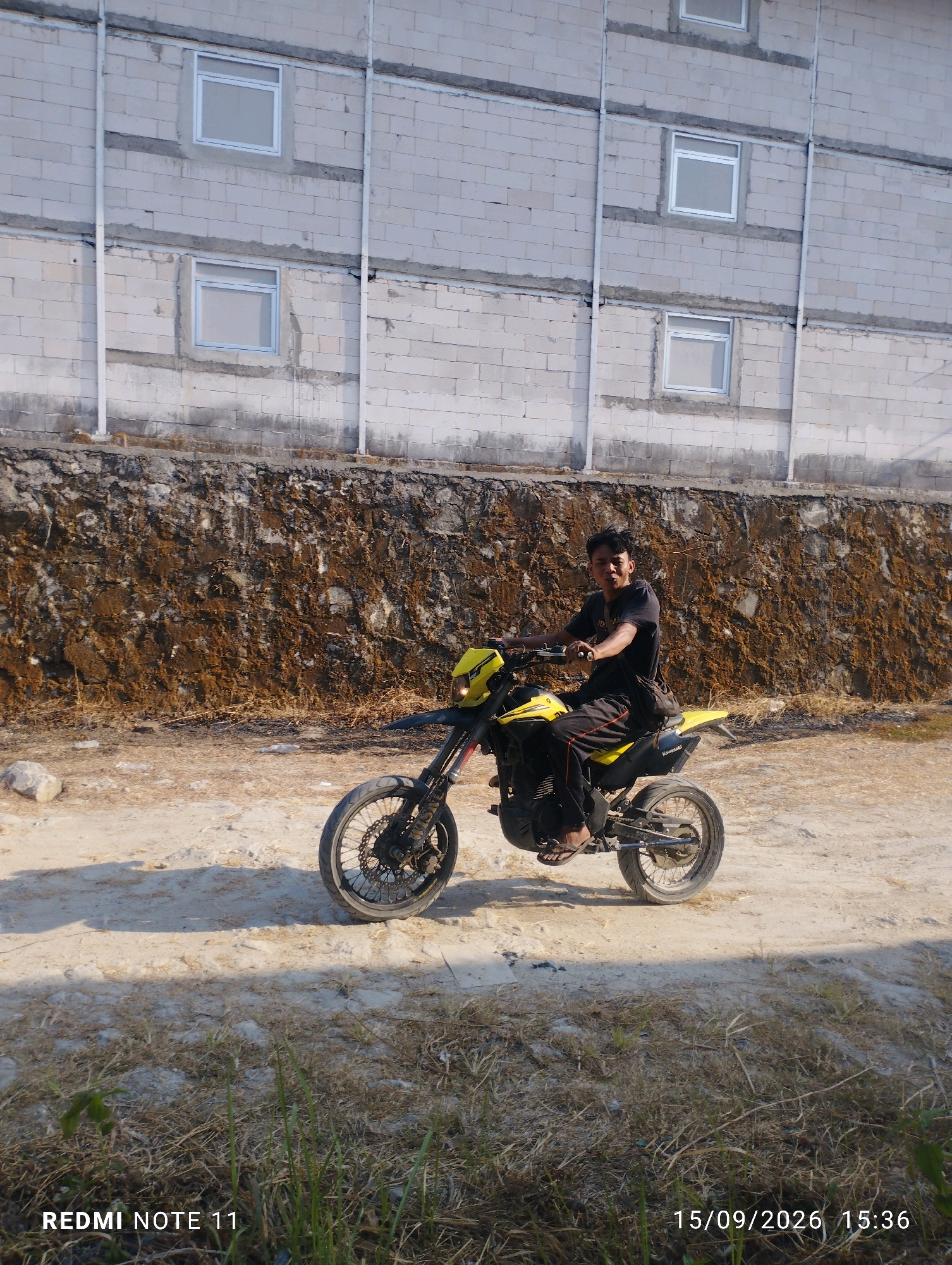 A young man sits on a yellow dirt bike, facing left. He is wearing a black t-shirt and black pants with a red stripe down the side, and appears to be looking at the camera. The bike has black tires, a black seat, and a black frame with yellow accents. In the background, there is a textured wall made of brown soil, with a building constructed of white bricks above it. The building has several windows with white frames. The ground in front of the bike is dirt and sand, with some dry grass and a few small rocks scattered about. The scene is illuminated by natural sunlight, casting shadows on the ground. The bottom right corner of the image shows text that reads "15/09/2026 15:36," and the bottom left corner reads "REDMI NOTE 11." This image was taken in Pulutan Wetan, Indonesia.