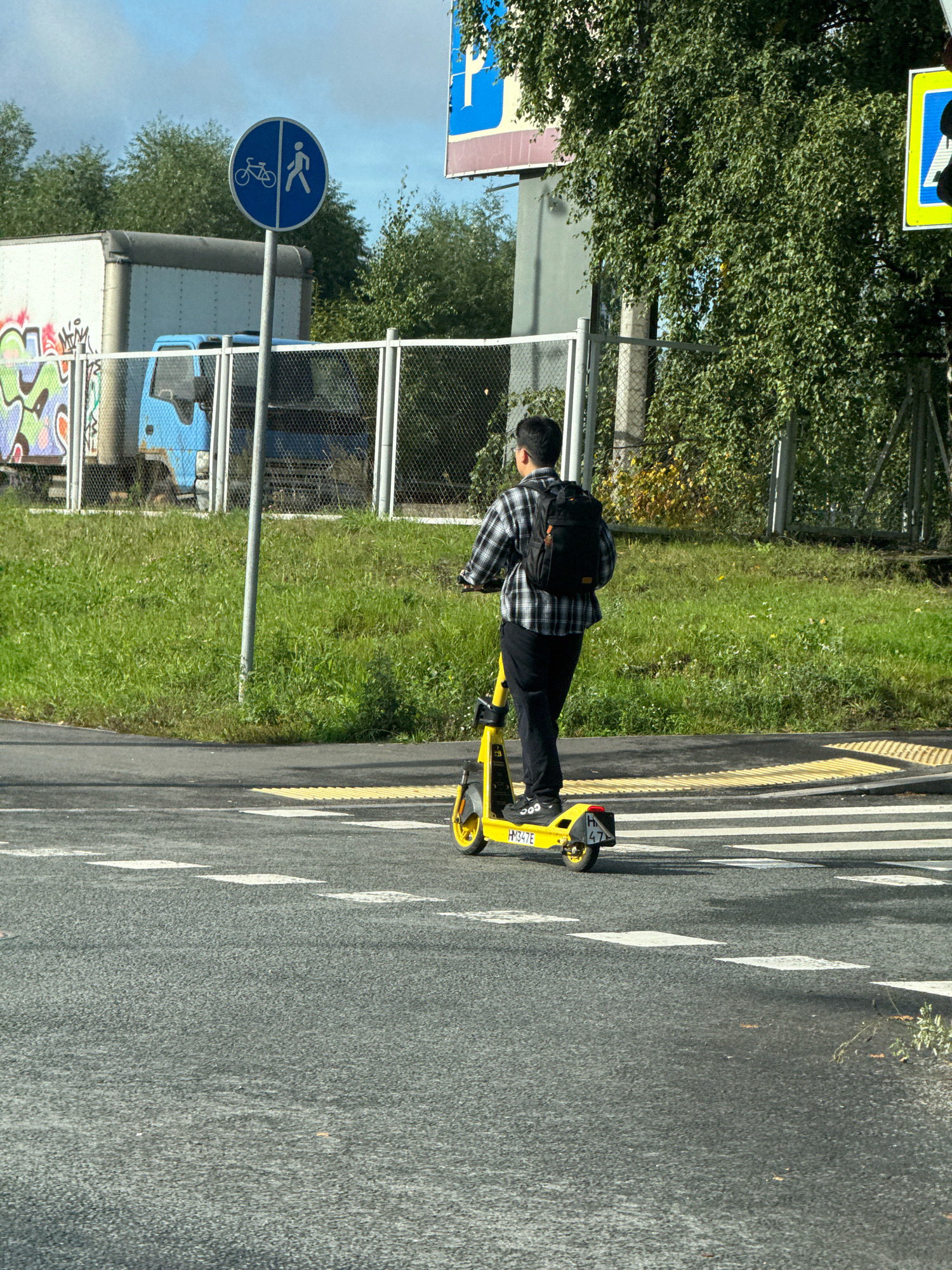 In an urban outdoor setting, a young man wearing a black and white plaid shirt, black pants, and a black backpack rides a yellow electric scooter on a paved road. He is crossing a pedestrian walkway indicated by white stripes. In the background, a blue truck with graffiti on its side is parked behind a metal fence. A traffic sign featuring a bicycle and a pedestrian is visible on the left, and trees and a cloudy sky complete the scene. The image appears to have been taken on a sunny day.