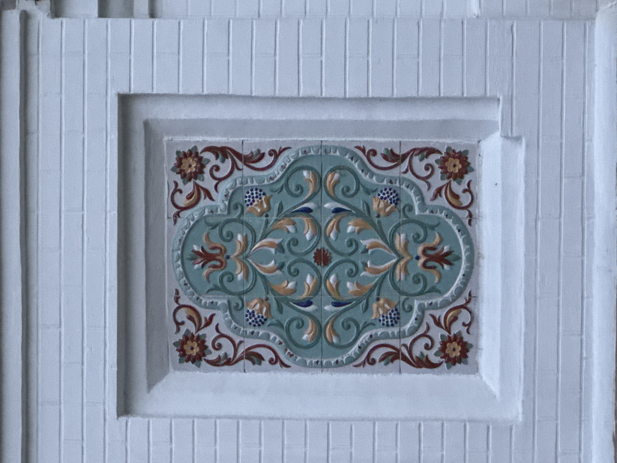 The image displays a close-up, eye-level view of a decorative tile panel set into a white, fluted surface. The tile panel features an intricate floral and scrolling design in shades of turquoise, deep red, gold, and blue. The central motif is an ornate, symmetrical pattern with stylized flowers, leaves, and circular elements. The outer border of the tilework is a lighter gray with red scrollwork and small, dark blue circular clusters that resemble berries or buds. The surrounding white surface is characterized by a series of vertical, parallel grooves, giving it a ribbed or fluted appearance. The lighting appears to be even and diffused, suggesting an indoor setting or an overcast day. There is no visible text in the image. The focus is entirely on the architectural detail and the decorative tilework, with no people or animals present. The overall impression is one of decorative craftsmanship.