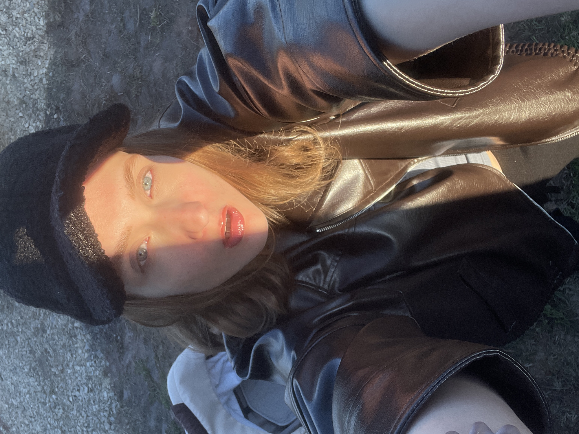 The image is a close-up, high-angle selfie of a young woman, taken outdoors in Podshivalovo, Russia. Her face is framed by light-colored hair and features striking blue eyes and glossy, red-tinted lips. She is wearing a dark, possibly faux fur, bucket hat and a shiny black faux leather jacket, with a white garment visible underneath. Strong, warm sunlight illuminates her face and the jacket, creating prominent highlights and suggesting either early morning or late afternoon. The background shows a gravelly or dirt ground interspersed with sparse green vegetation. The photo captures a casual moment with a focus on her expression and stylish attire. No text is visible.