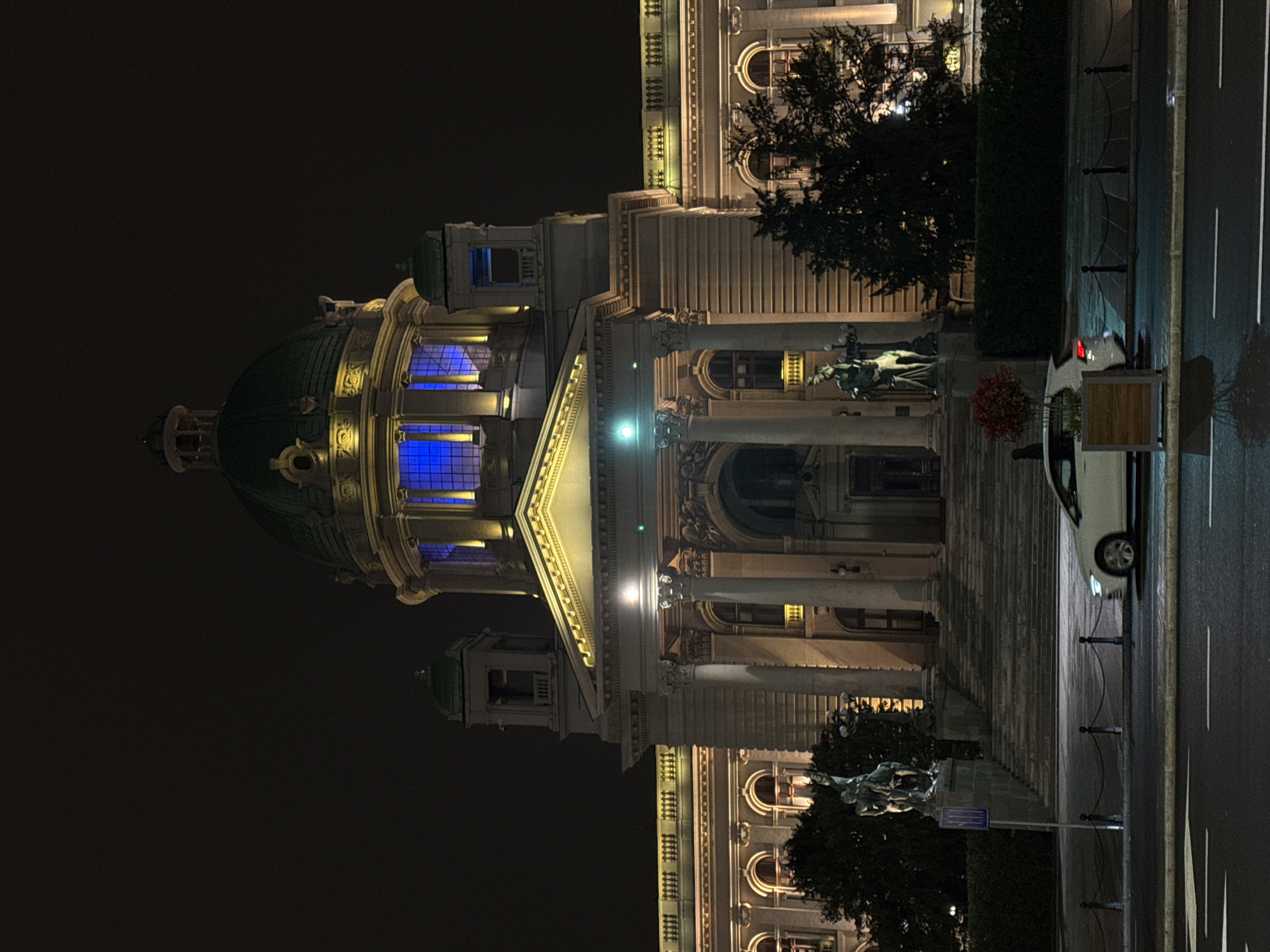 Night view of the National Assembly of Serbia in Belgrade. The building has a green dome and is illuminated with yellow and blue lights. A statue stands in front of the building. A car is parked alongside the street, and trees are adjacent to the building.