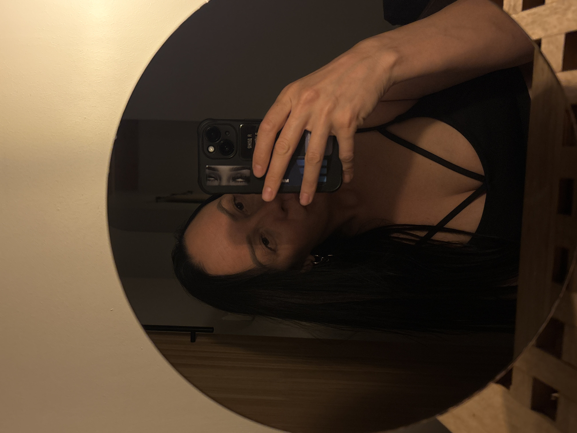 The image shows a person taking a selfie in a round mirror. The person is holding a black smartphone in their right hand, obscuring part of their face. The phone case has a sticker with a pair of eyes on it. The person has long, dark hair and is wearing a black top with strap details across the chest. The background is blurry but appears to be a room with light-colored walls and wooden furniture. There is no evidence suggesting the location is Chaikovsky, Russia.