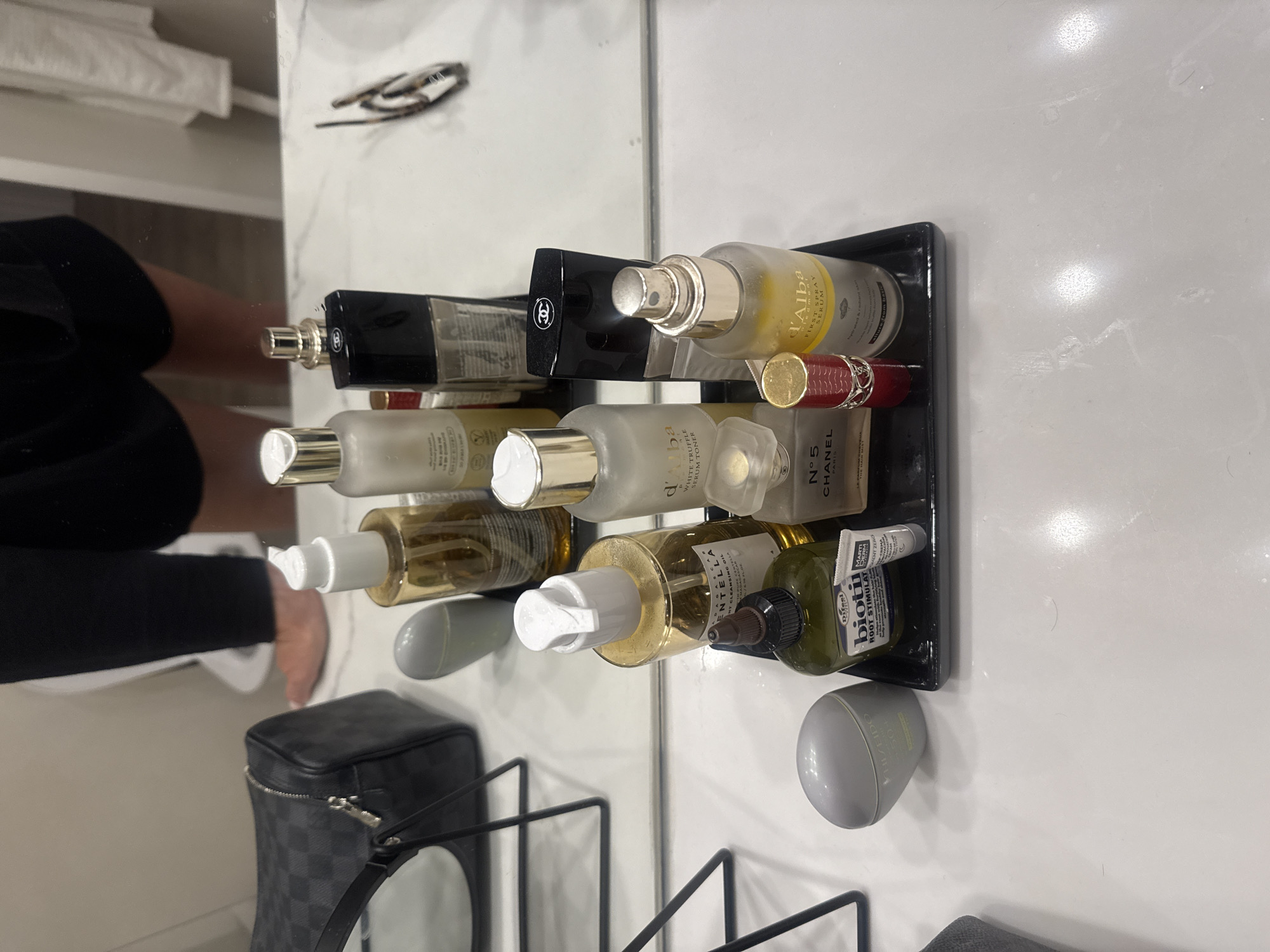 The image shows a variety of beauty products arranged on a black tray, which is placed on a white surface that appears to be a bathroom countertop. Products include: Chanel No. 5 perfume, d'Alba First Spray Serum, d'Alba White Truffle Serum Toner, A'pieu Pure Source Snail Cream, and Biotin Root Stimulant. Additionally, a gray cosmetic bag is nearby, along with an unidentified gray container. The location is not discernible from the image. The image shows a cluttered or disorganized bathroom environment. No people or actions are distinctly captured in the main area of focus. The mirror in the image reflects a person's leg and arm, suggesting someone is present but not directly involved in the scene of the beauty products.