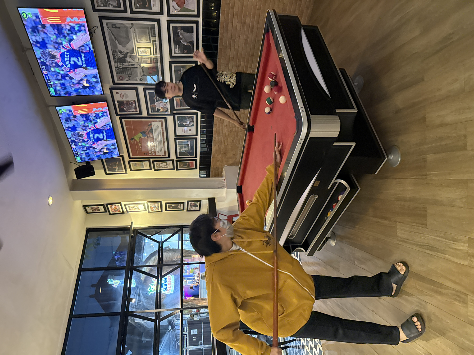 This image depicts an indoor recreational area, likely a bar or game lounge in Kuta, Indonesia, where two young men are playing billiards. One individual, dressed in a yellow hoodie, black pants, and black sandals, is positioned on the left side of the red-felt pool table, holding a cue and wearing a face mask. His attention is focused on the game. Opposite him, another young man, wearing a black t-shirt with a distinctive white bat or wing graphic and black pants, is poised over the table, also holding a cue and preparing to strike. Several billiard balls are scattered across the table's surface.
The walls surrounding the area are extensively decorated with numerous framed pictures and posters, many featuring sports legends and events, with visible names like "NADAL" and "SAMPRAS," as well as "CANO SYD 1T" and "EW 3". Above the pool table, two wall-mounted flat-screen televisions are displaying a sports match, possibly rugby, featuring a player wearing jersey number "2." On-screen text includes "FOX," "UNIBET," and a partial McDonald's logo ("Ma"), along with game scores and timers such as "0:0:59" and "3:10." A team name fragment "Crsteck" is also visible.
Large windows on the left side of the room reflect the indoor lighting and an external neon sign that reads "SLUSH ISLAND," suggesting the scene takes place during the evening or night. The room features a light-colored wooden floor. The overall atmosphere is one of casual recreation and entertainment.