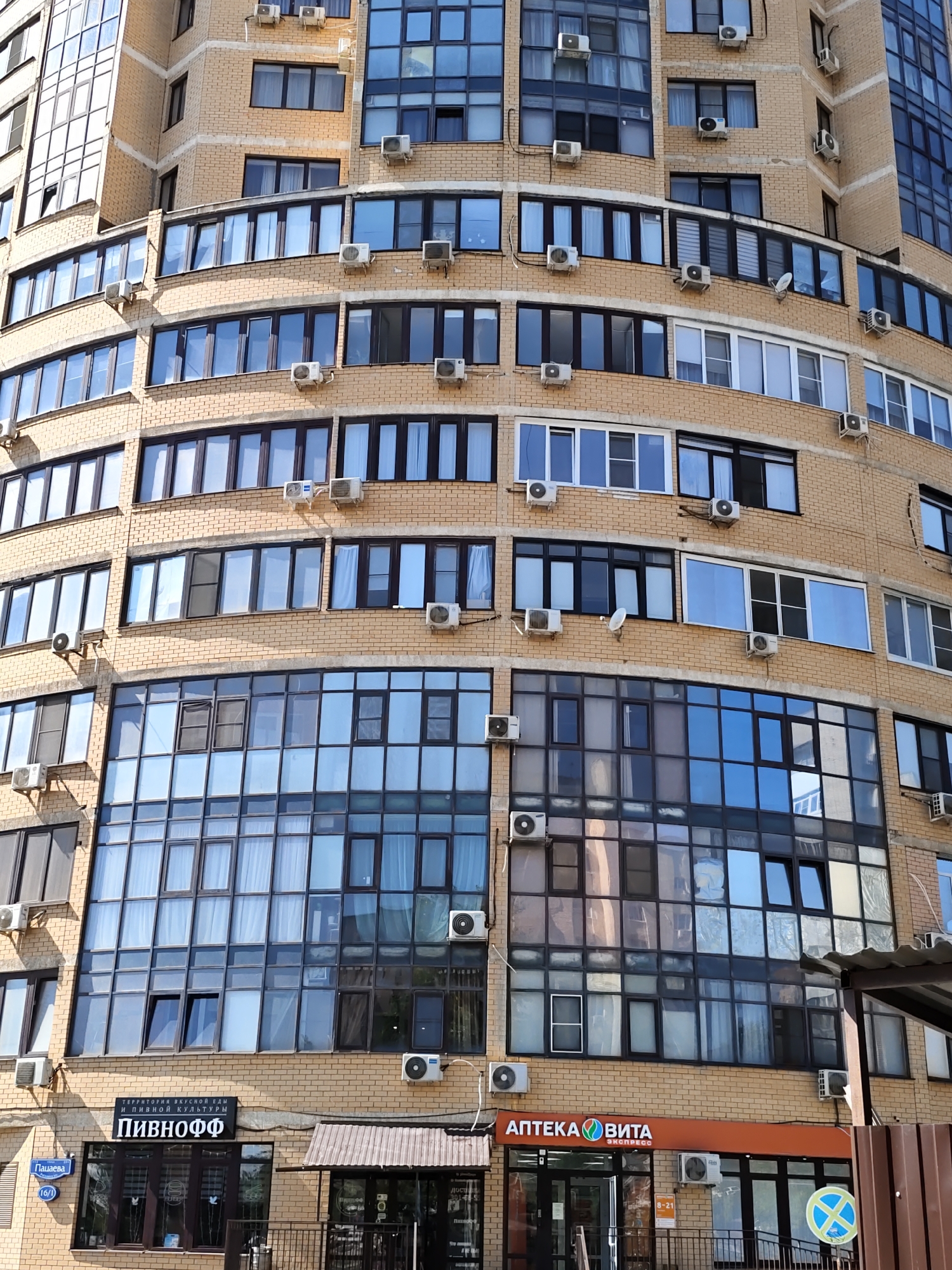 The image shows the facade of a modern, multi-story building in Rostov-on-Don, Russia. The building is constructed with light brown brick and features numerous windows, many of which are large and panoramic, reflecting the sky. Several air conditioning units are visible mounted on the exterior walls.
At street level, there are commercial establishments. One storefront has a sign with Russian text that includes "ПИВНОФФ" and "ТЕРРИТОРИЯ ВКУСНОЙ ЕДЫ И ПИВНОЙ КУЛЬТУРЫ". Another storefront has a prominent orange sign with Russian text "АПТЕКА ВИТА ЭКСПРЕСС". A street sign with Russian text "Пацаева 16/1" is visible on the left side of the building. The weather appears to be clear and sunny. There are no people or animals visible in the image.