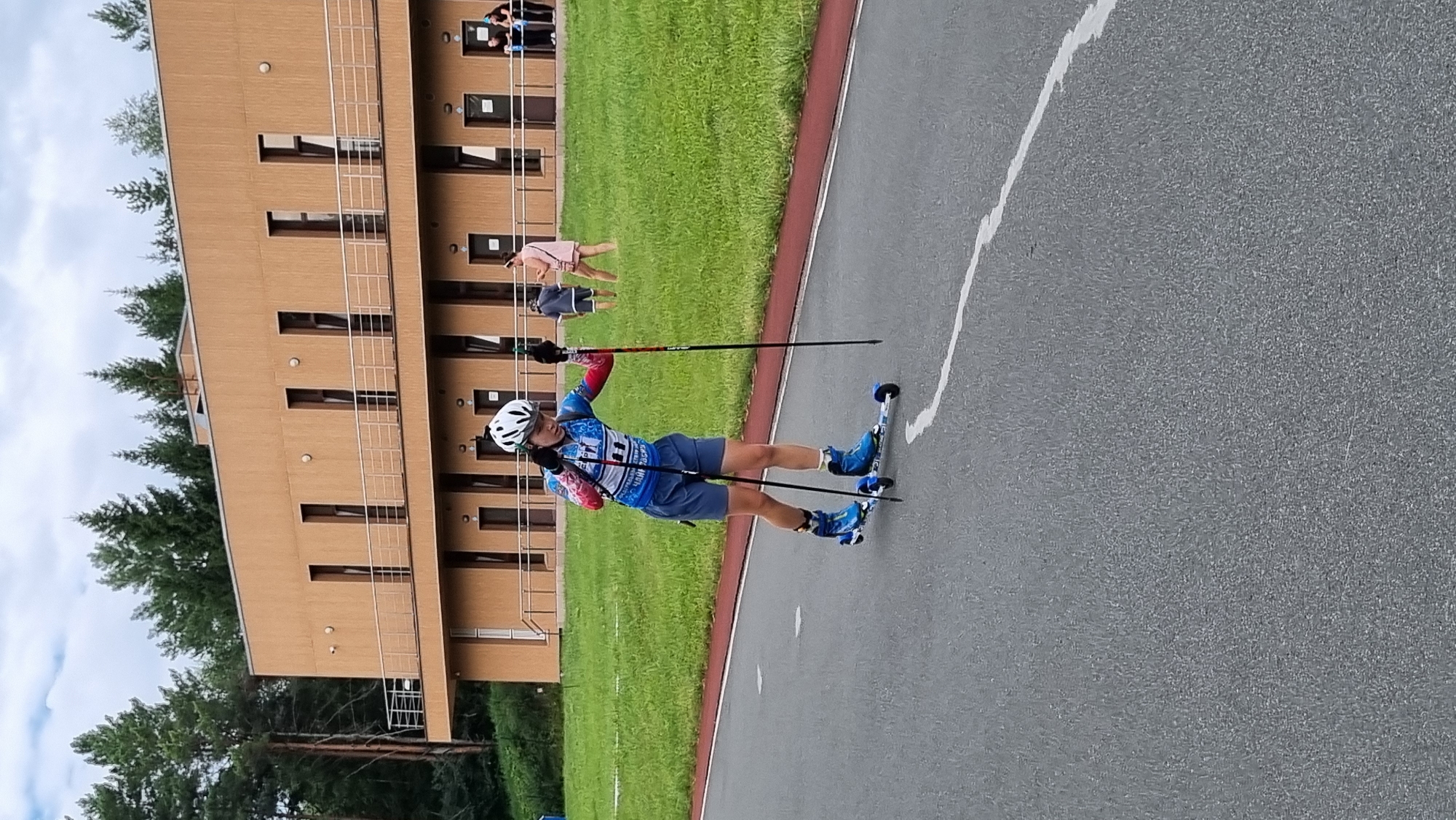 A young female athlete, wearing a white helmet, blue and red athletic top, and blue shorts, is roller skiing on an asphalt track. She is holding two poles, one in each hand, which are black with red tips. Her roller skis are blue and have three wheels each. The track is dark gray with a red border and a white curved line on the right side. In the background, there is a tan building with multiple windows and a balcony. Behind the building, there are tall green pine trees and a cloudy sky. Several other people are visible in the background, some standing near the building and others walking on the grass. The scene appears to be an outdoor sports facility, possibly located in Прикамский, Russia, given the context.
