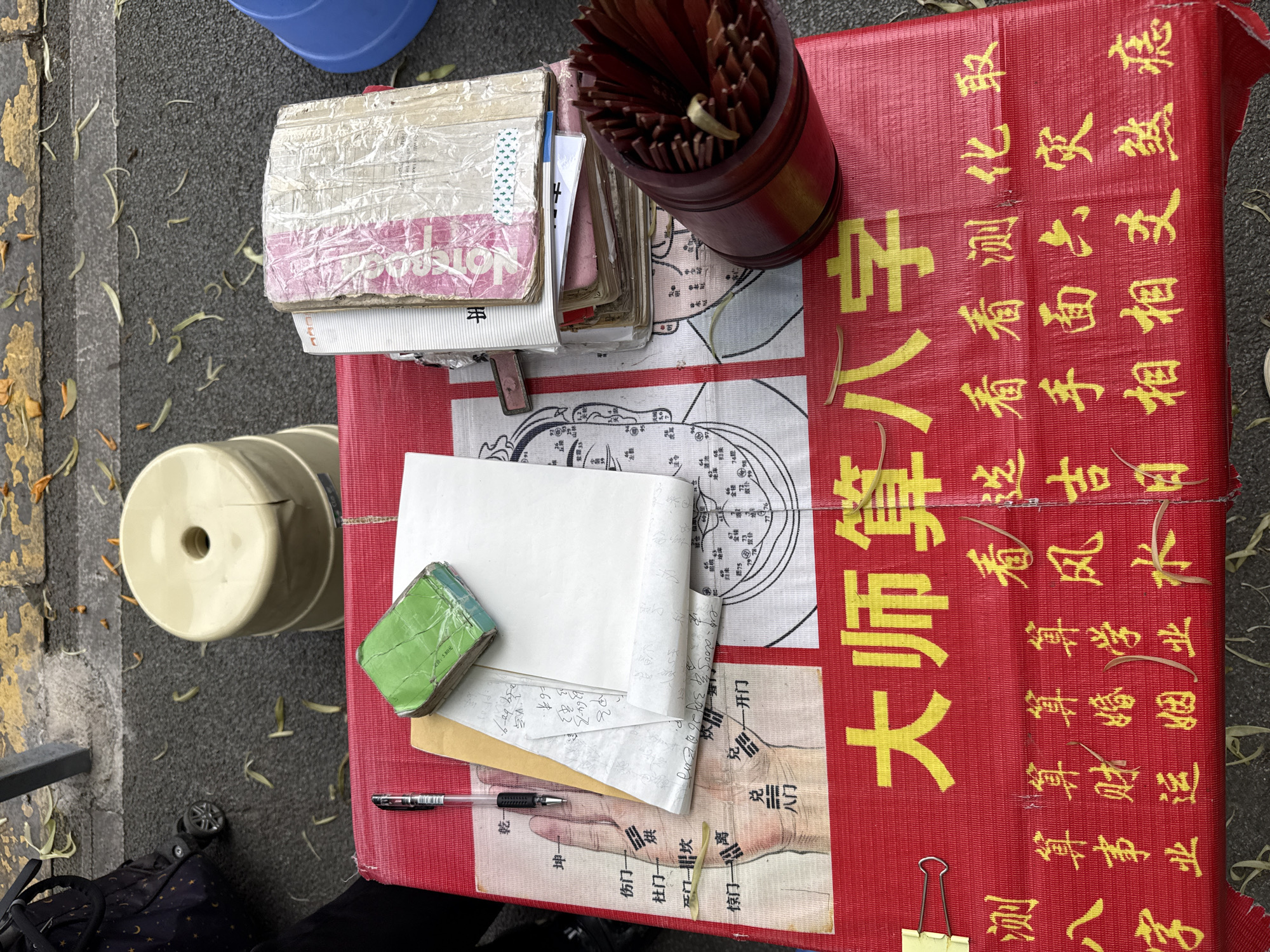 The image depicts a fortune teller's setup on a street in Chongqing High-tech Zone, China. A red cloth with Chinese characters, likely advertising fortune-telling services such as birth charts, career, wealth, and marriage predictions, covers a surface. Various fortune-telling paraphernalia are scattered across the cloth. These include stacks of books, a cup filled with divination sticks, a green inkpad, blank paper, a pen, and diagrams of hands and acupressure points. A yellow clip is visible. Stools are placed near the setup. A glimpse of a person's legs and a patterned bag is visible at the bottom. The street surface shows signs of wear and is littered with small debris, including what appears to be fallen leaves or petals.