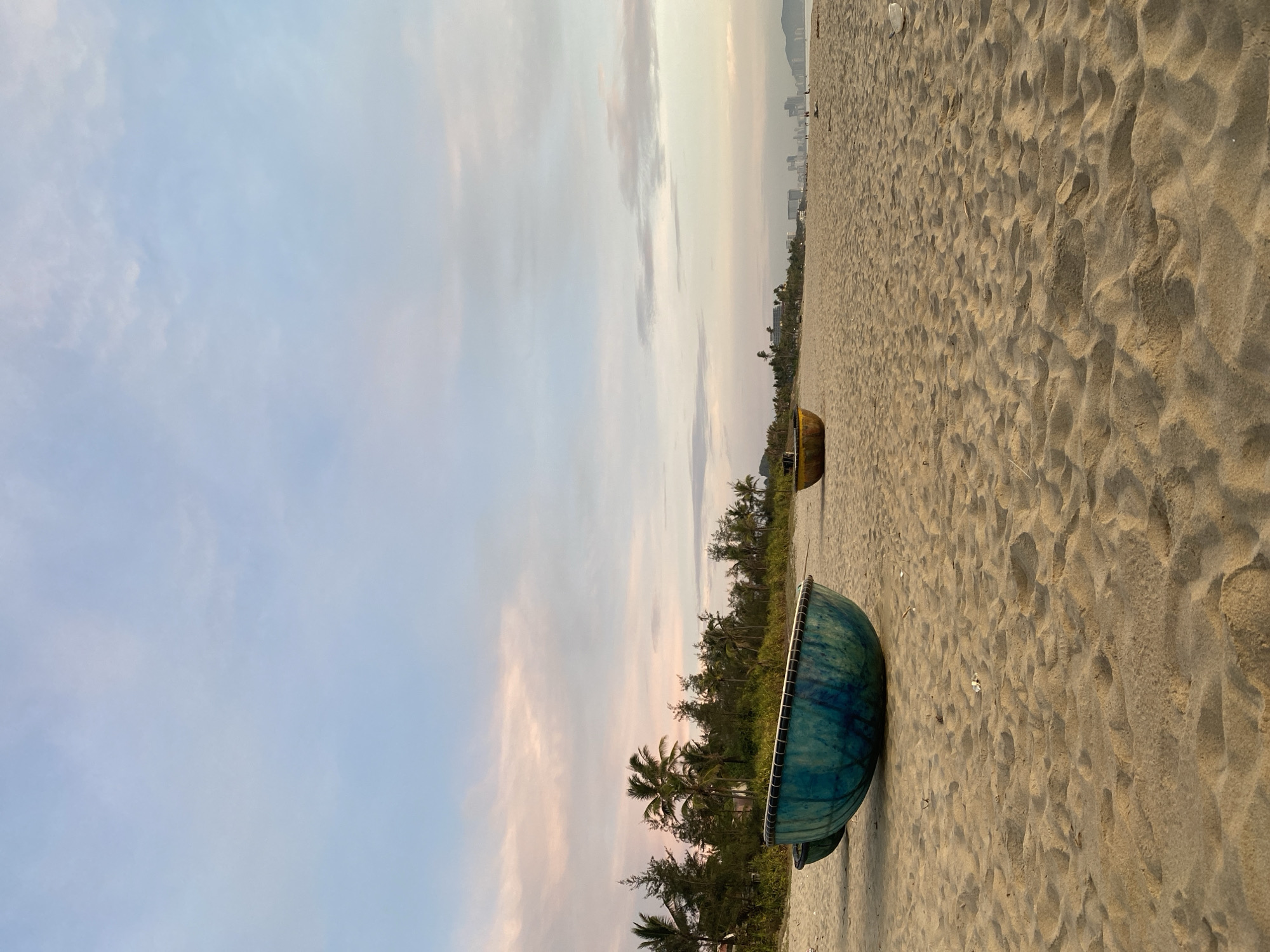 The image depicts a tranquil beach scene at dawn or dusk. The sand is a light tan, with visible footprints and textured undulations suggesting a gentle breeze. In the foreground, a large, teal-colored coracle-shaped boat rests on the sand, its rim lined with white rope. To the right and further back, a smaller, yellow coracle boat is visible.
The background features a line of lush green palm trees and other tropical foliage, hinting at a coastal location. Beyond the trees, a cityscape with numerous buildings, including high-rises, is faintly visible on the horizon, suggesting an urbanized area. The sky is a soft blend of pale blue and pink hues, with scattered clouds, indicative of either sunrise or sunset. A few distant figures can be seen walking on the beach in the far distance.
This scene is characteristic of My Khe Beach in Đà Nẵng, Vietnam, known for its long stretches of sand, palm-lined shores, and proximity to the city.