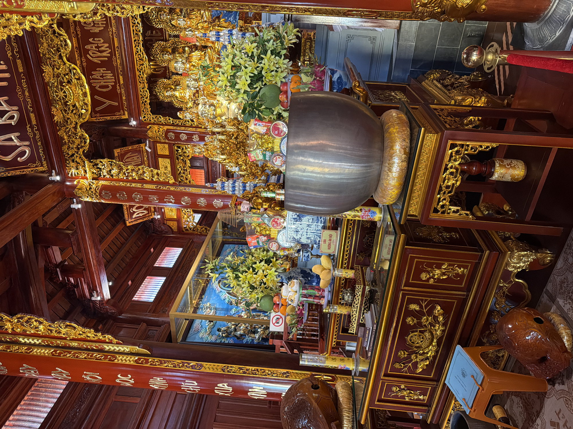 This is an interior shot of a temple in Diên Khánh, Vietnam. The location is identifiable by the text on the walls. The temple features dark wooden architecture with elaborate gold detailing. There are various shrines or altars holding offerings, including a large metal bowl filled with flowers and fruit. A glass display case holds more offerings and religious artifacts. The scene is well-lit and suggests a place of worship or reverence. An orange plastic stool can also be seen.