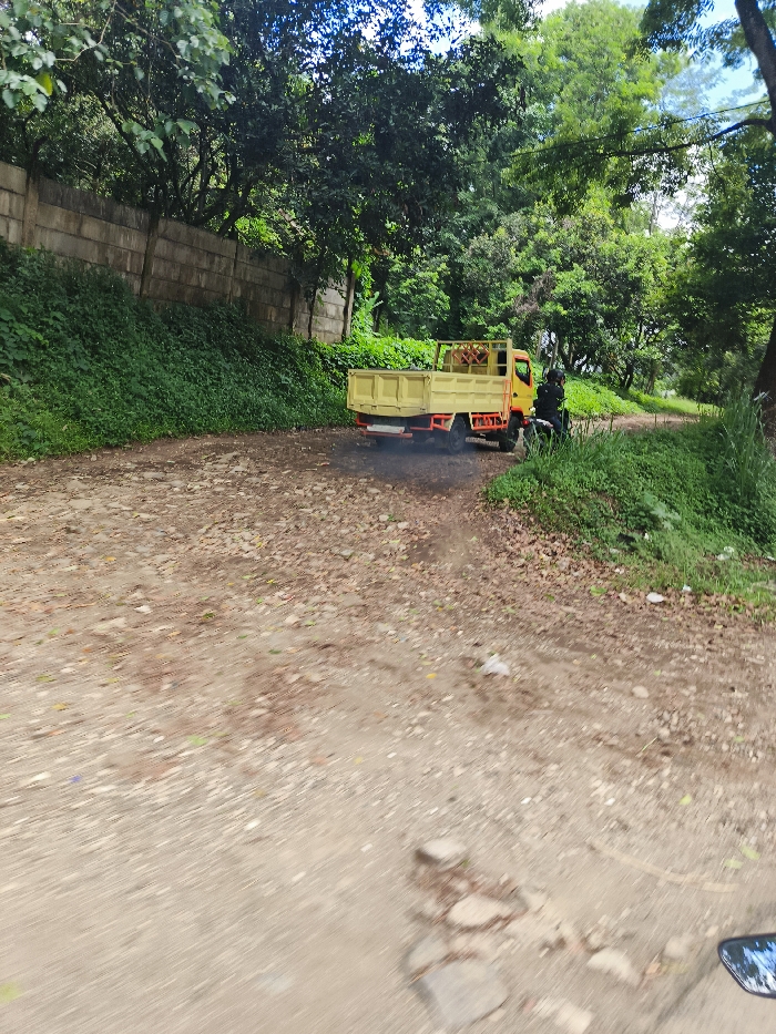 A yellow truck is driving down a dirt road in the town of Cipatat, Indonesia. The truck is carrying an unknown load in the back. The road is flanked by vegetation. There is a motorcycle parked on the right side of the road. The motorcycle's rearview mirror is reflecting a blurry image. The truck is driving away from the camera. The coordinates provided are in a rural part of Cipatat. The coordinates are 107.442° E longitude and -6.796° S latitude. The image is likely captured from a moving vehicle looking out the rear window.