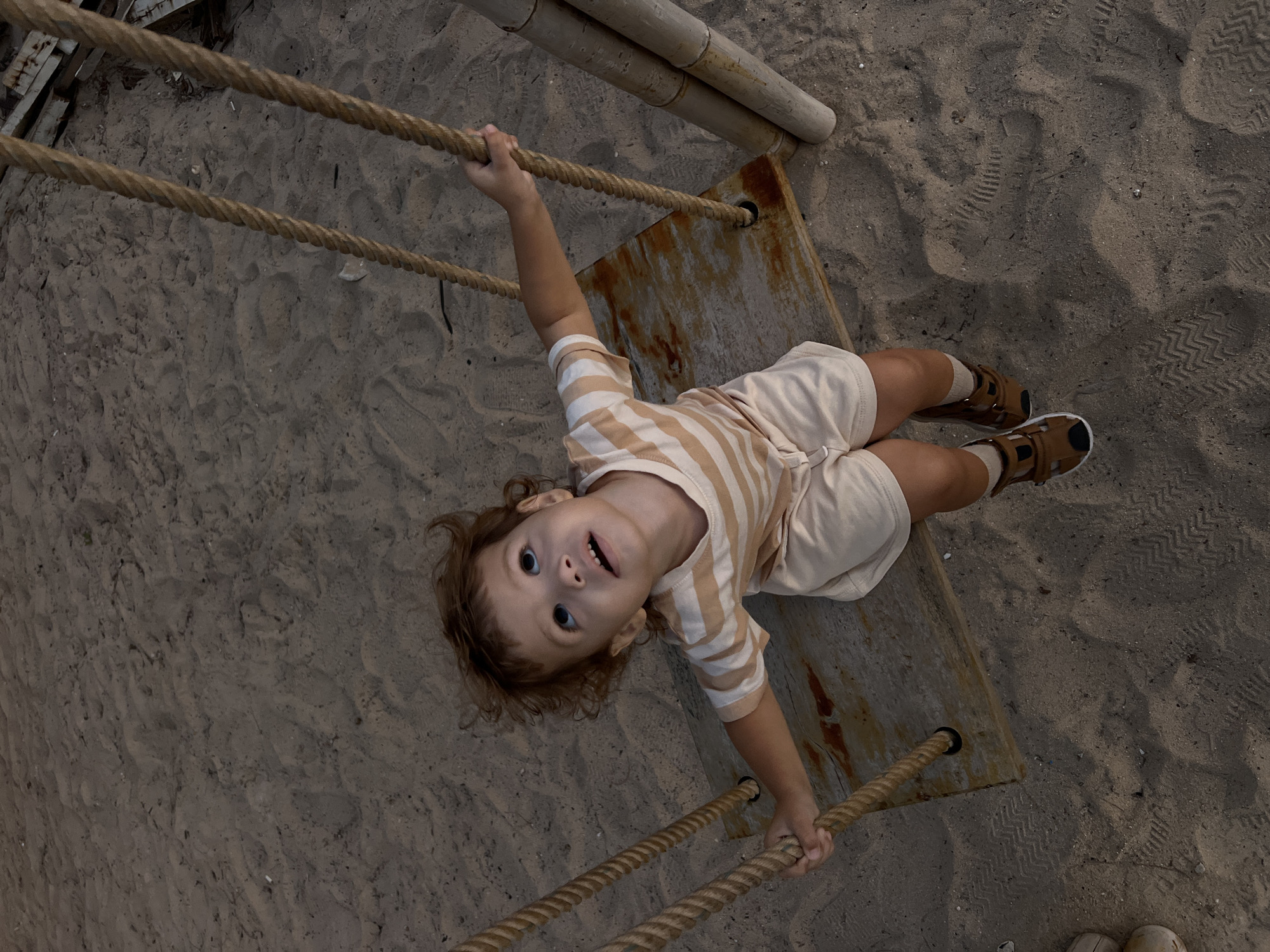 A high-angle, medium shot captures a young child with curly reddish-brown hair sitting on a wooden swing with thick rope supports. The child is looking upwards with an open mouth, appearing excited or surprised. They are wearing a light-colored t-shirt with horizontal tan stripes and matching tan shorts, along with tan sandals and socks. The swing is suspended over a sandy surface that shows tire tracks and footprints. To the right of the swing, two large bamboo poles are partially visible. The scene appears to be outdoors, possibly a beach or playground, with the lighting suggesting it is either early morning or late evening. The location is identified as Choeng Thale, Thailand.