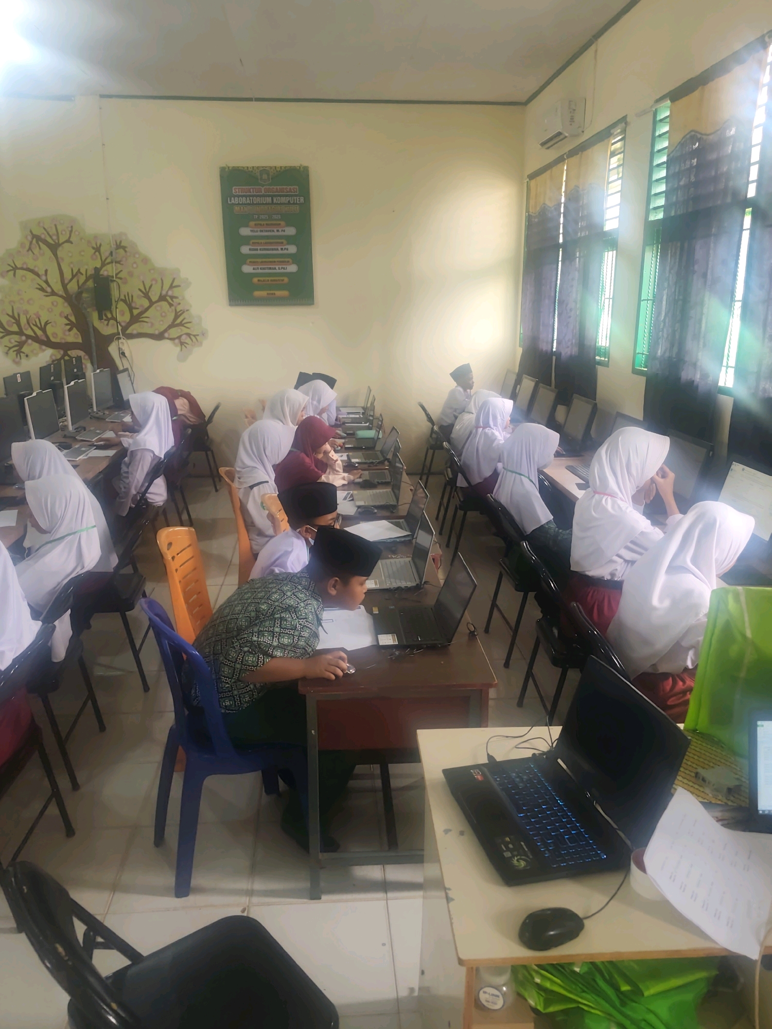 The image depicts an indoor scene of a computer lab, likely at a school in Pematang Reba, Indonesia. Several young students, predominantly wearing white hijabs and school uniforms, are seated at desks, engaged with computers. Some are looking intently at their screens, while others appear to be typing or interacting with the devices. The room has a light-colored wall with a painted mural of a tree on the left side. A poster with text, likely an organizational chart for the computer lab, is displayed on the wall. The lab is equipped with multiple computer monitors and laptops arranged on desks. Natural light streams in from windows covered with curtains on the right side of the room. The flooring appears to be tiled. The overall atmosphere suggests a classroom setting where students are participating in a computer lesson or activity.