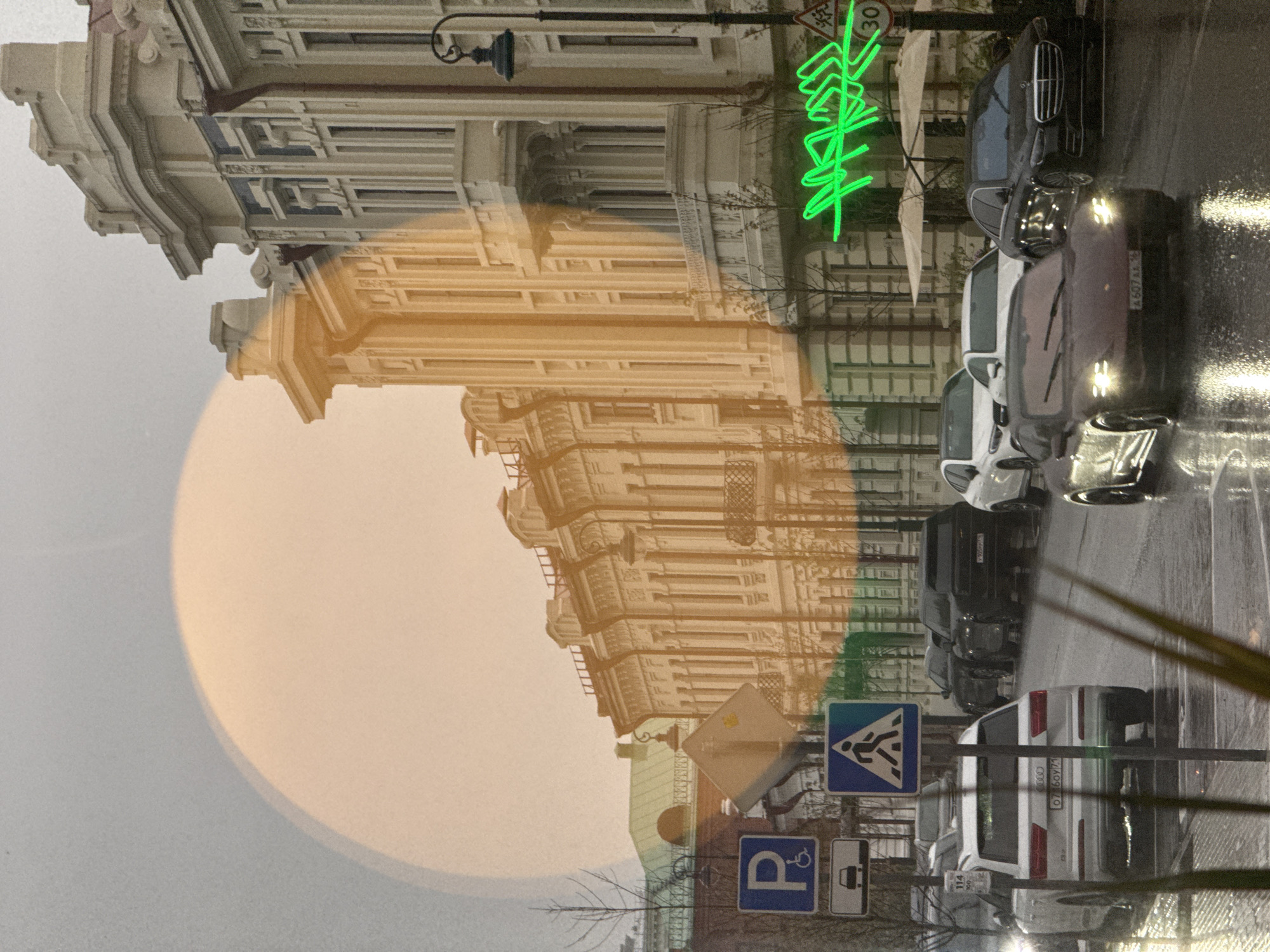 The image depicts a street scene in Kazan, Russia, with parked cars lined along a wet street. A building with ornate architectural details is visible in the background, overlaid with an orange, circular shape. A green neon sign is affixed to a building. Street signs for parking and pedestrian crossings are present. The image is oriented vertically. The environment appears to be overcast or rainy.