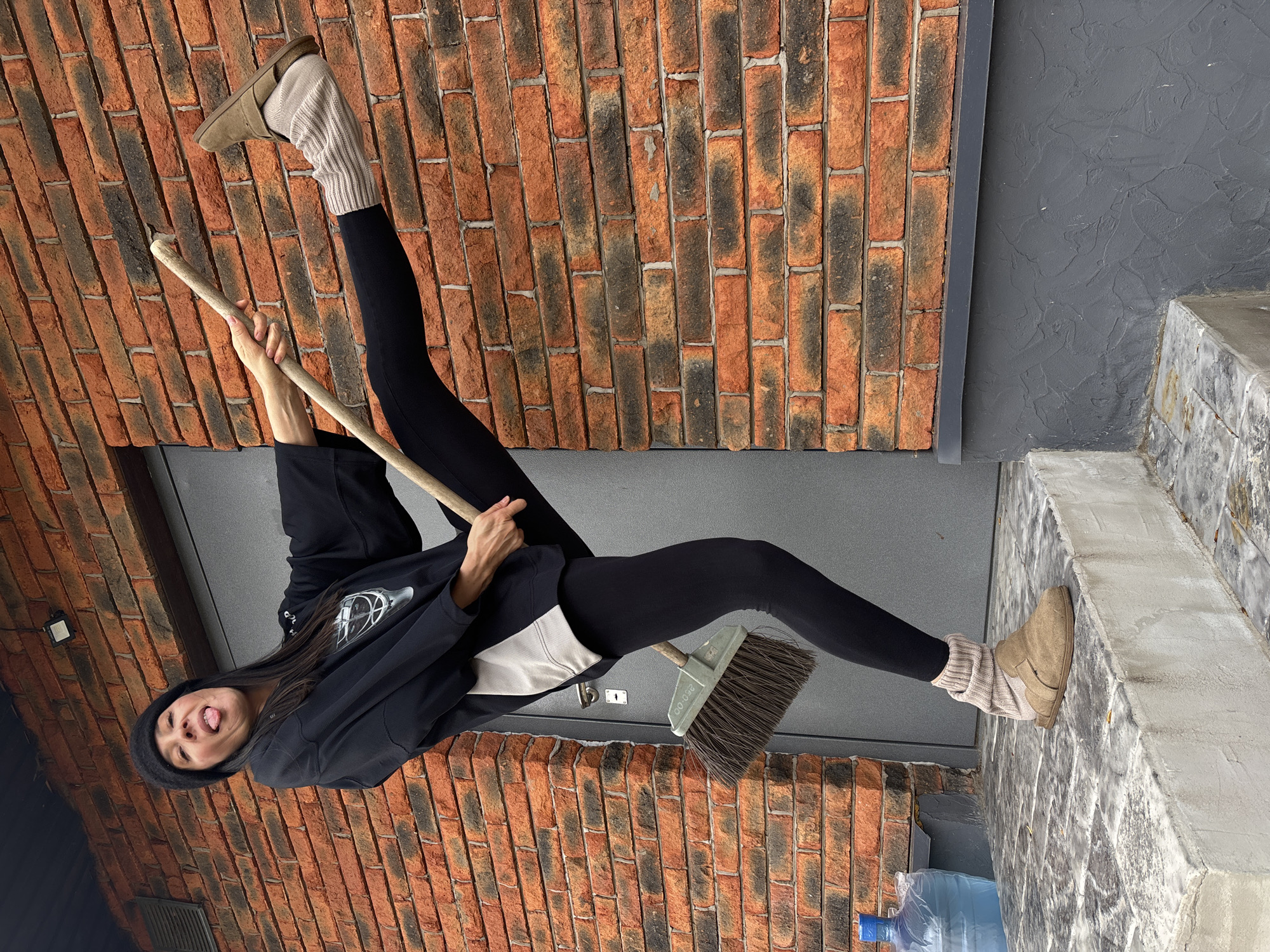 A young woman is captured in a dynamic and playful pose against a brick and dark gray wall background. She is depicted seemingly "flying" or elevated, holding a long wooden stick with both hands as if sweeping, with a broom head positioned near her bent right leg. Her left leg is extended upwards. She is dressed in a black beanie, a black hooded sweatshirt over a lighter top, black leggings, and light brown slip-on shoes paired with ribbed beige leg warmers. Her facial expression is cheerful, with her tongue sticking out towards the viewer, conveying a sense of humor and energy.
The background consists of a prominent brick wall, featuring a mix of orange-red and darker, burnt-looking bricks. To the right, a dark gray, textured wall or door panel is visible, next to a concrete ledge or step. A large clear plastic water bottle with a blue cap rests on the ground in the lower left corner. The lighting appears even, suggesting it's daytime, possibly in a sheltered outdoor area or indoors. Text "250.00" is visible on the broom head. The overall scene gives the impression of a creative and unconventional photo opportunity. The image was taken in Chaikovsky, Russia.