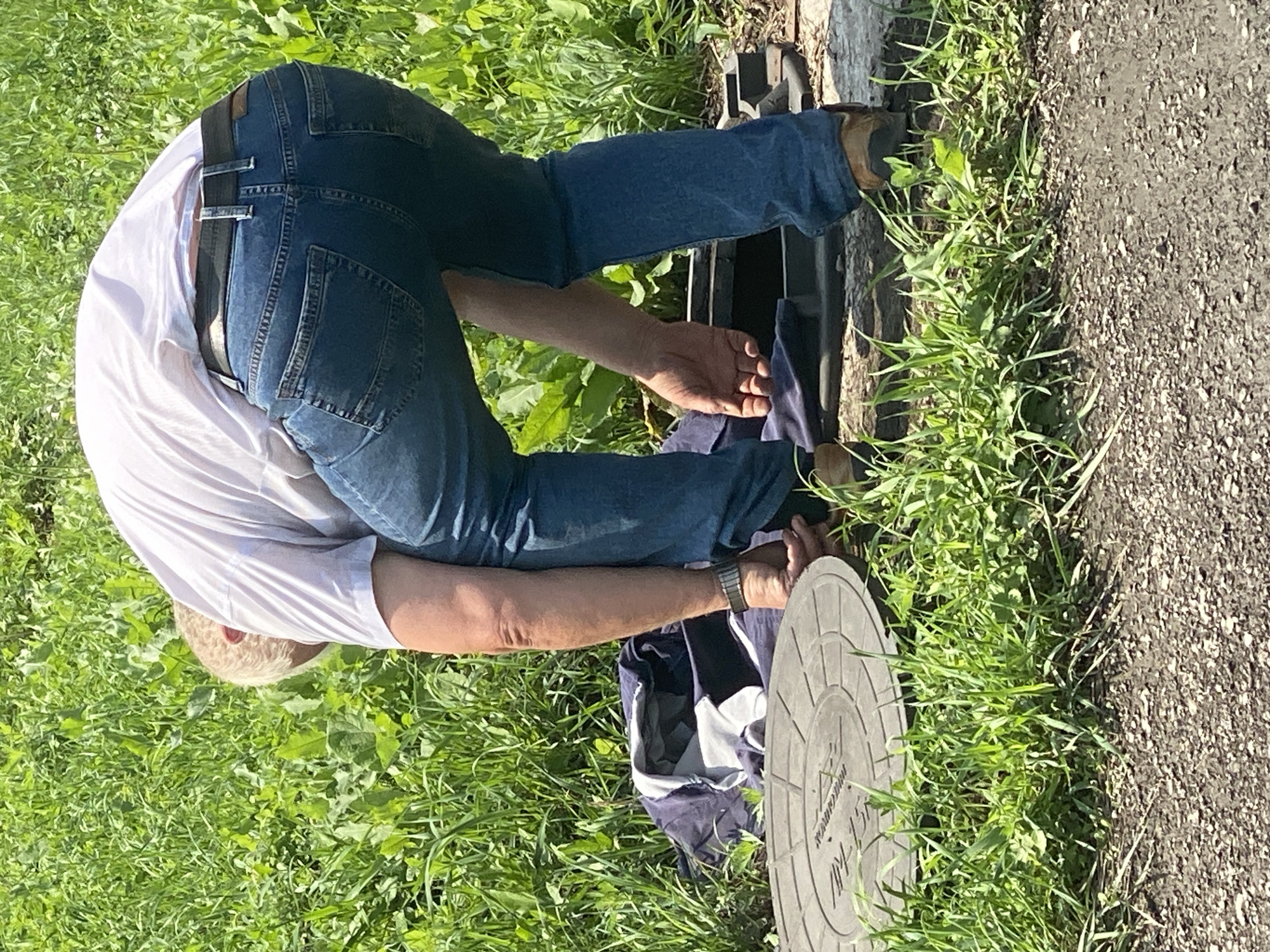 A man in a white t-shirt and blue jeans is bending over in a grassy area next to a gravel path. He appears to be interacting with a manhole cover and some items that have been placed on the ground. One item is a dark blue and white jacket or bag. Another is a circular, dark-colored object with a raised pattern, possibly a lid. The man is wearing a watch on his left wrist. The environment suggests an outdoor urban or semi-urban setting with vegetation. The location is identified as Novokuznetsk, Russia.