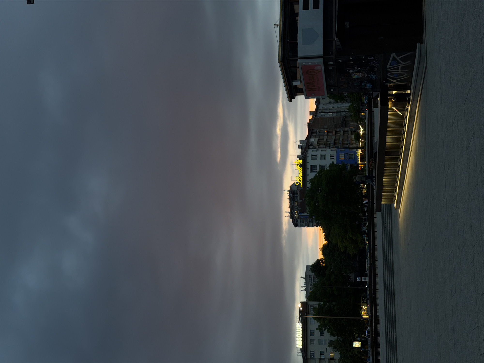 The image shows an evening scene in Sofia, Bulgaria, likely near the National Palace of Culture (NDK). The sky occupies the left side of the image, displaying a dark, overcast sky with a hint of sunset colors along the horizon. A series of buildings stretches horizontally across the middle of the frame, interspersed with trees. There are neon signs visible, including one that reads "ABSOLUTE." On the right side is a concrete stairway with built-in lights along its steps. Graffiti is visible on the sides of the stairway. The image suggests a moment just before dusk in an urban environment.