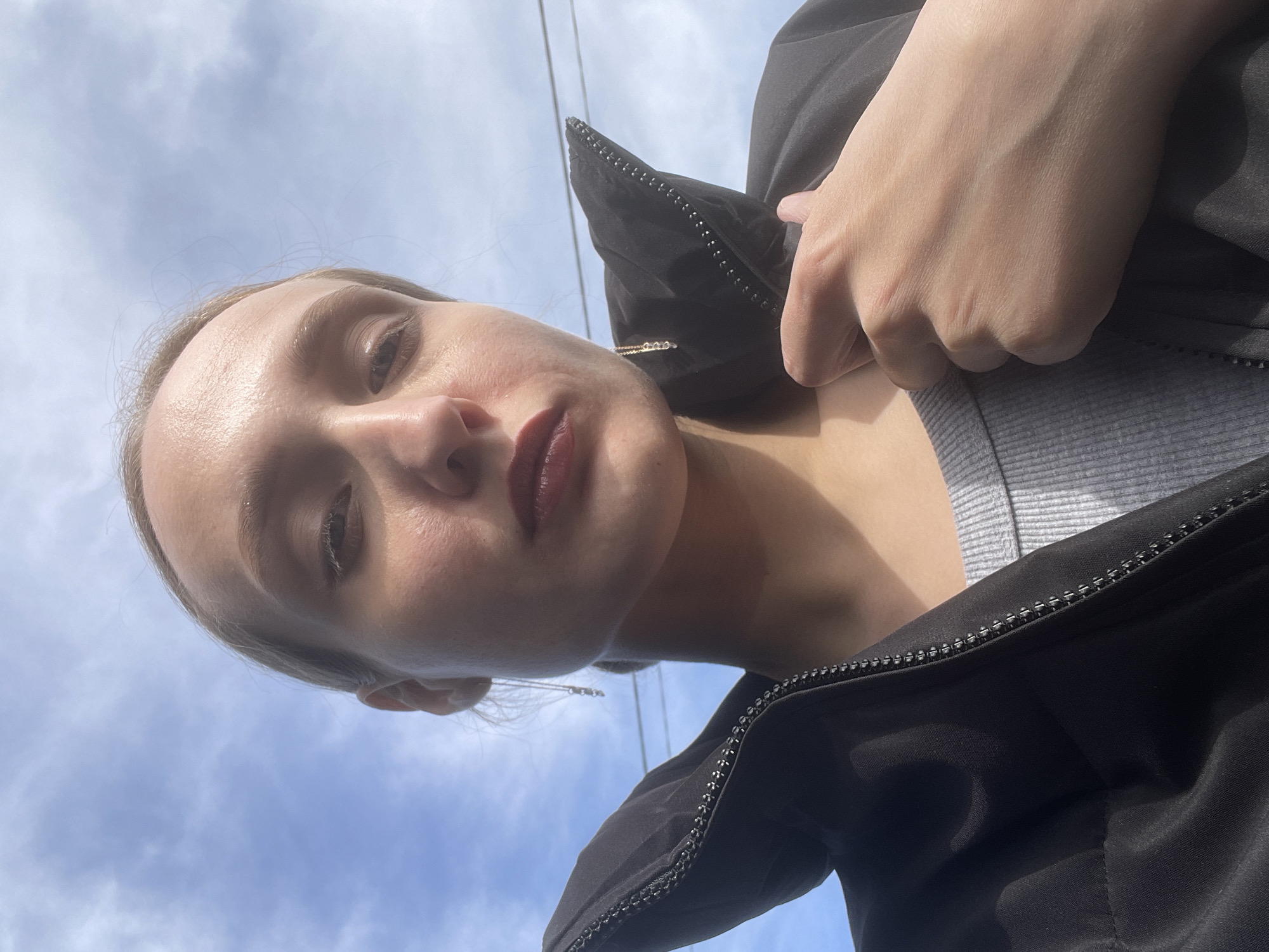 A woman is taking a selfie. She is wearing a black jacket over a grey shirt. The background shows a blue sky with some white clouds. The picture was taken from a low angle, looking up at the woman. It is not possible to confirm the location of Podshivalovo, Russia, from the image alone.