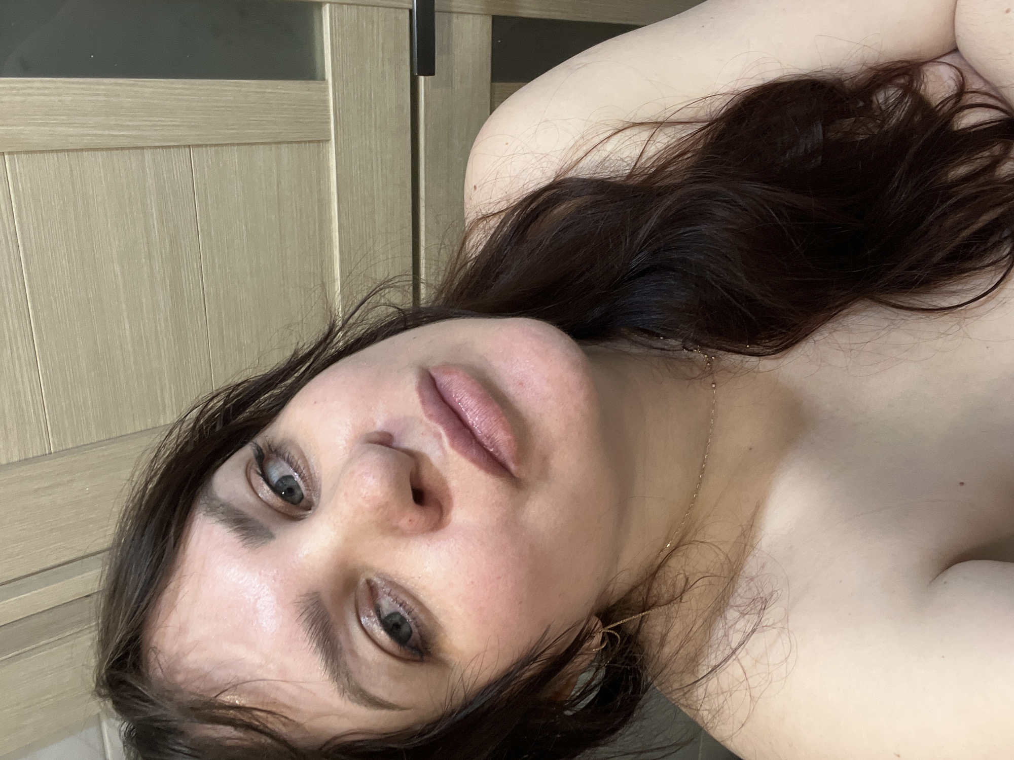 A close-up, angled shot features a woman with dark hair and light skin. She is wearing a thin gold necklace. Her eyes are visible, and her lips are a soft pink color. The background includes a light-colored door with a dark handle. It is not possible to verify the location of Noviy, Russia, from this image.
