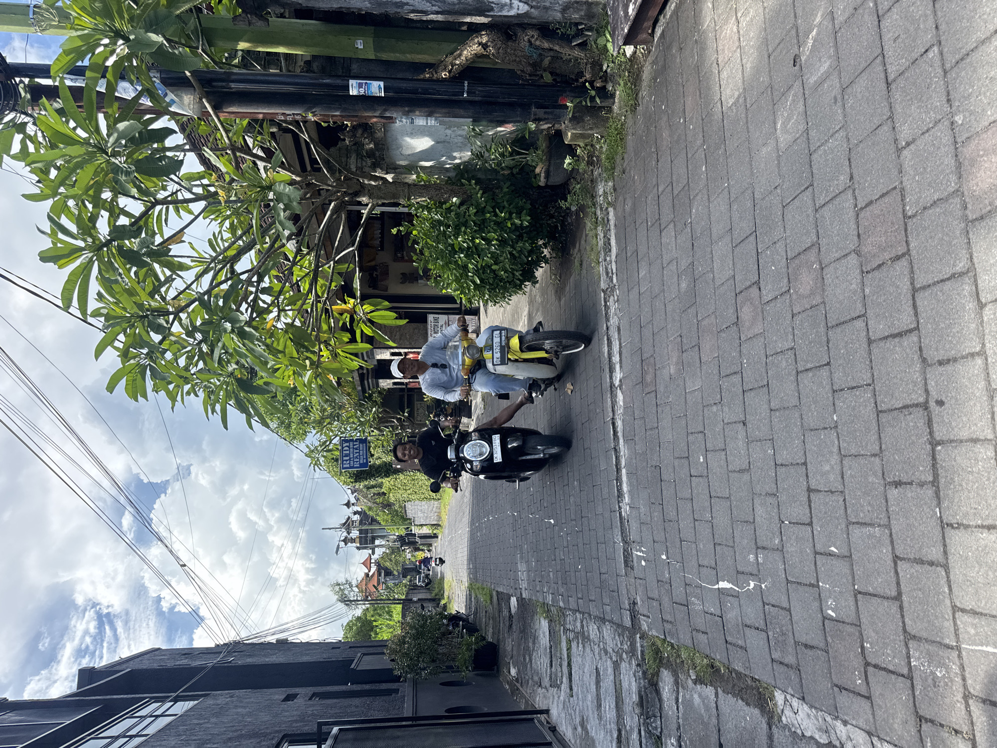 The image depicts a narrow street scene in Kuta, Indonesia, characterized by grey brick pavement and bordered by buildings and lush tropical foliage. Two individuals are riding motor scooters down the street. One rider, closer to the foreground, is on a black scooter and wears a black shirt. The second rider, positioned further down the street, is on a yellow and white scooter, clad in a light-colored shirt and a white hat. A business sign labeled "Rudy Rental" is visible, positioned perpendicular to the street. Overhead, a network of electrical or communication wires stretches across the sky, which is partly obscured by the buildings and trees. The sky exhibits patchy cloud cover. Buildings on either side of the street have varying architectural styles, with one side showing a more modern structure and the other older, more traditional Balinese-style buildings. Vegetation including trees and shrubs is evident on both sides of the street. Other scooters are parked in the distance.