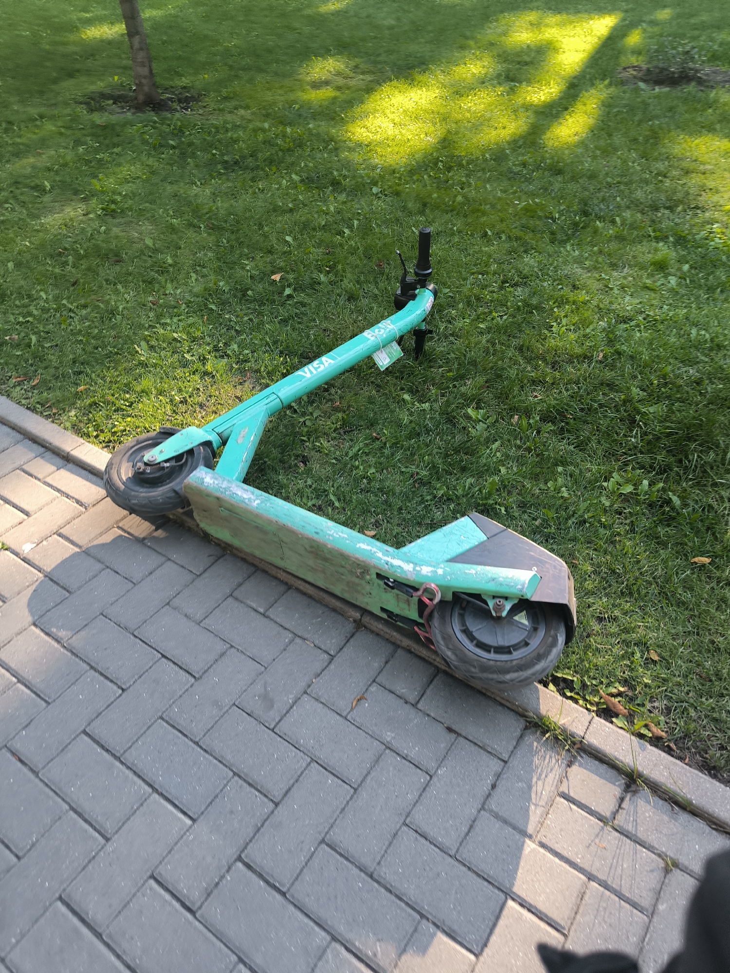 An electric scooter, painted teal and white with the "Bolt" logo, lies on its side. The scooter is positioned on a paved walkway next to a grassy area, with the handlebars and front wheel extending onto the grass. The scooter appears to be abandoned or unattended. The environment suggests a public space, possibly a park or sidewalk in Chișinău, Moldova, given the context of the prompt. The lighting indicates it is daytime, with shadows cast across the scene, suggesting the sun is at an angle. The ground is a mix of grey brick pavers and green grass. No people are visible in the image.
