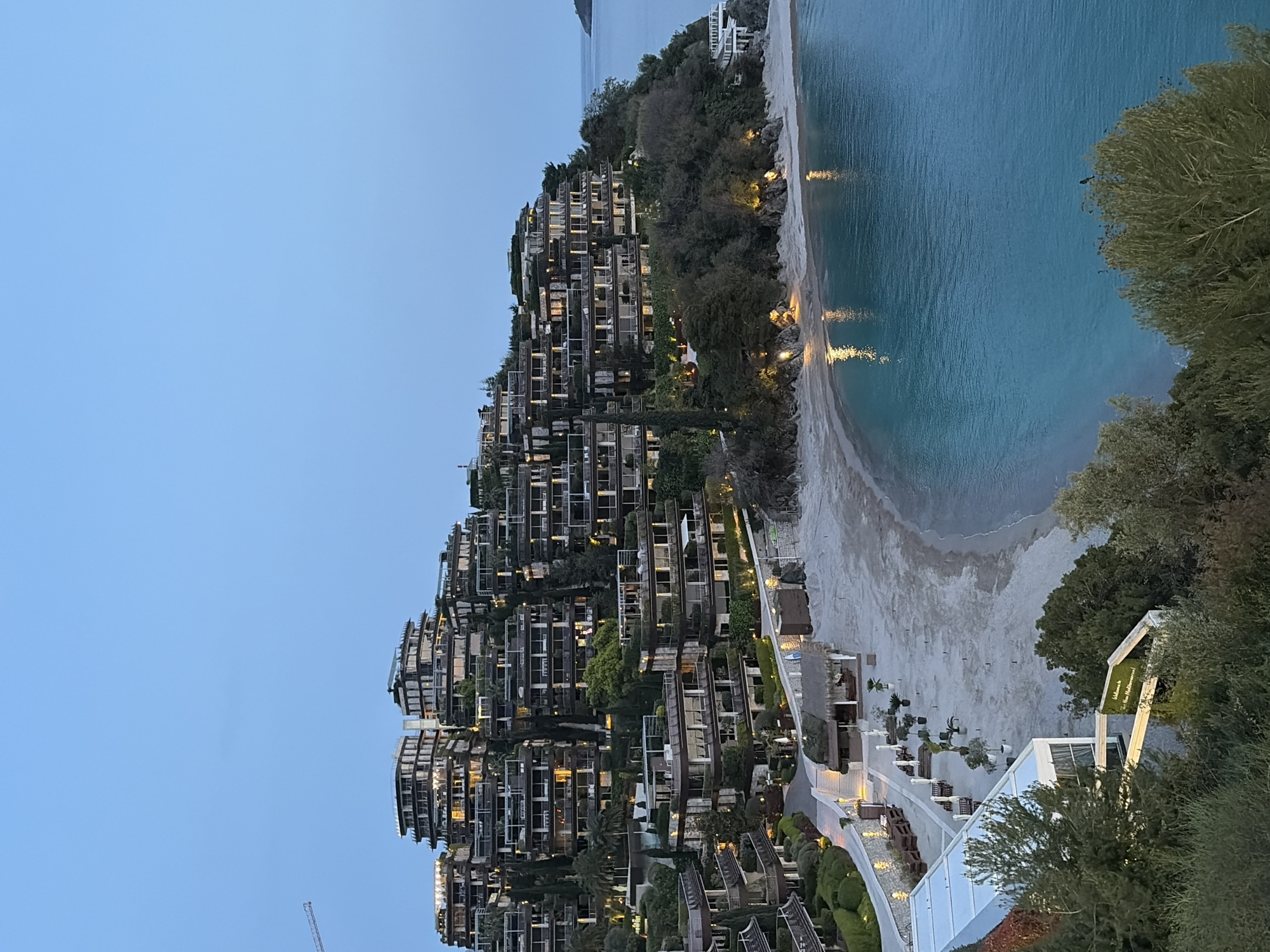 The image depicts a coastal scene in Budva, Montenegro, featuring a hillside development of modern buildings cascading down towards a beach. The buildings are characterized by a terraced design with multiple levels, likely apartments or residences. The architecture incorporates glass and metal elements, with numerous balconies and illuminated windows. Lush greenery is interspersed throughout the development, integrating the buildings into the natural landscape.
The sandy beach curves along the coastline, bordered by turquoise waters. Reflections of lights are visible in the water near the shore. A portion of the beach area is developed with walkways, landscaping, and possible resort or recreational facilities. In the lower right corner, architectural details and landscaping are visible, including outdoor seating areas and manicured hedges.
The sky is a pale blue, suggesting either early morning or late evening light. The scene suggests a high-end residential or resort development in a scenic coastal setting.