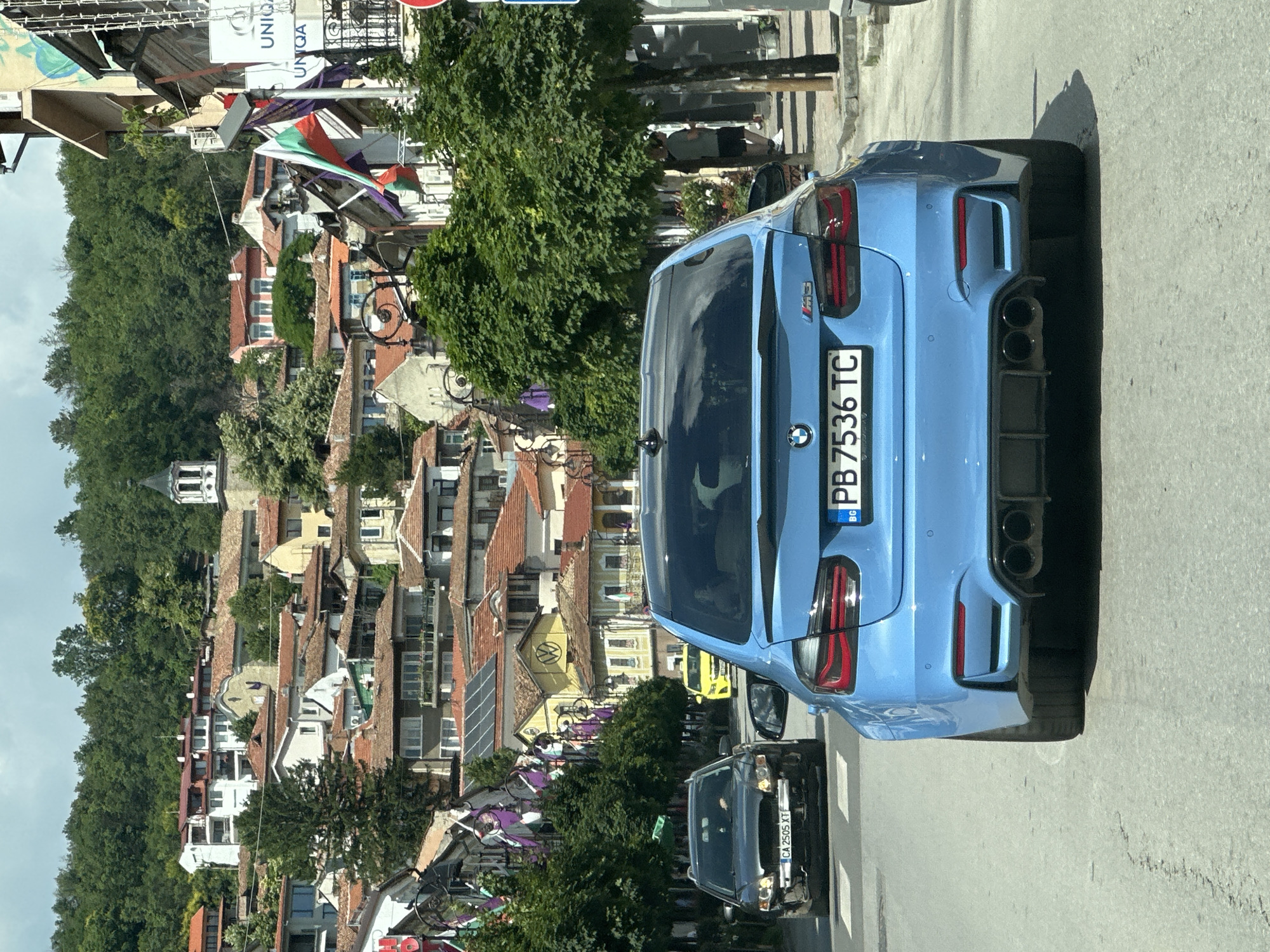 The image depicts a view from a vehicle driving down a street in Veliko Tarnovo, Bulgaria. The street is lined with traditional Bulgarian houses with red-tiled roofs, characteristic of the city. Green trees and vegetation cover the hillsides behind the buildings. On the right side of the street, there are shops and a Bulgarian flag is prominently displayed.
In the foreground, a blue BMW M3 is driving on the road. In front of it, a dark-colored car, possibly a Volvo, is also driving. Further ahead, a yellow taxi is visible. The sky is cloudy. The license plate of the blue BMW is visible and reads "PB 7536 TC". The license plate of the dark car reads "CA 2505 XT".
