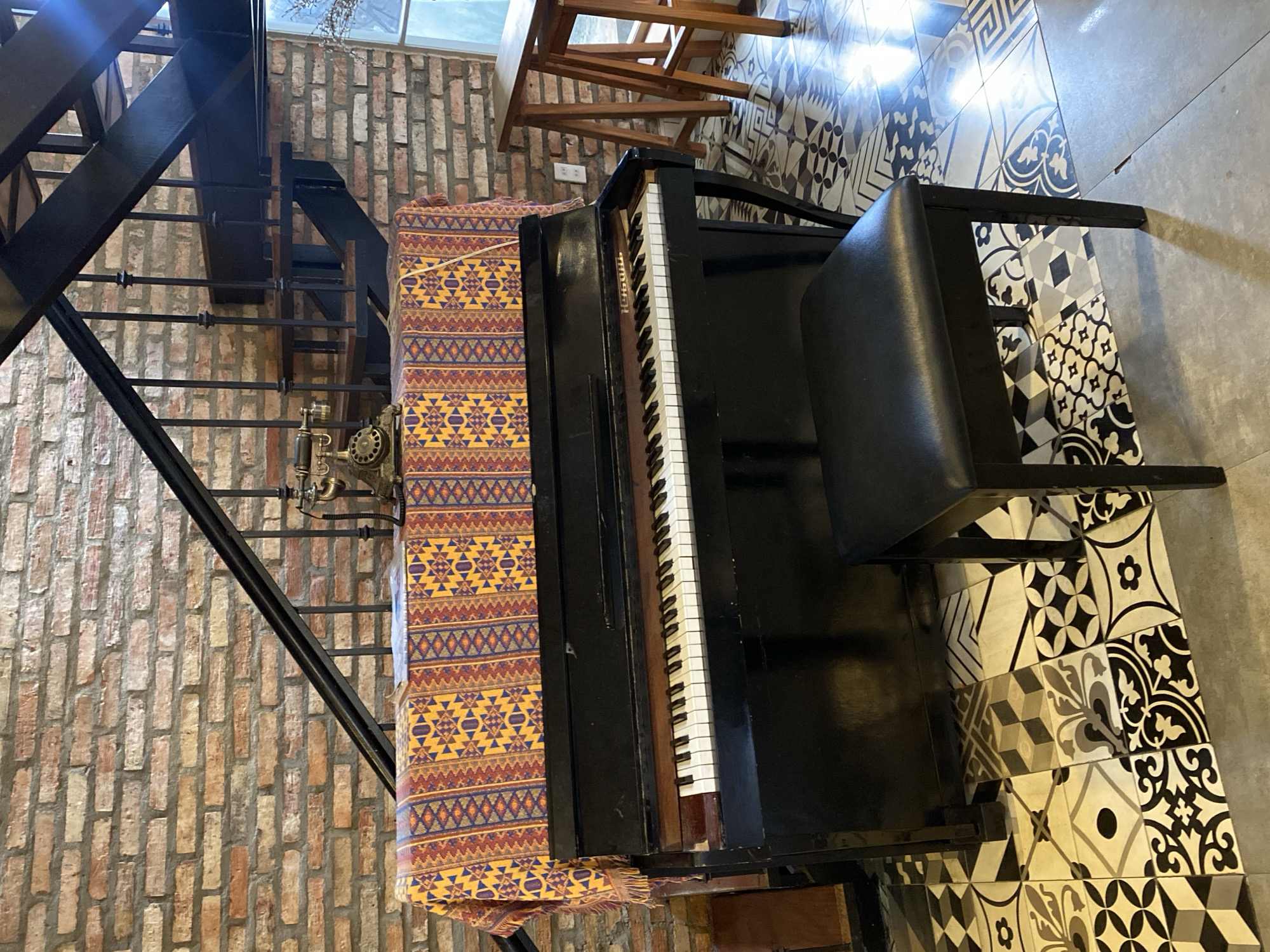 A vintage upright piano with a black finish is positioned in front of a brick wall. A colorful, patterned tablecloth is draped over the top of the piano, and an antique telephone rests on it. A black piano bench with a padded seat is placed in front of the piano. The floor is covered with decorative tiles in various geometric and floral patterns. A black metal staircase with wooden steps ascends behind the piano. In the background, a wooden table with a minimalist design stands near a window. The scene appears to be set in a cafe or a home with a rustic and vintage aesthetic, likely in Da Lat, Vietnam, known for its French colonial architecture and bohemian vibe. There are no people visible in the image.