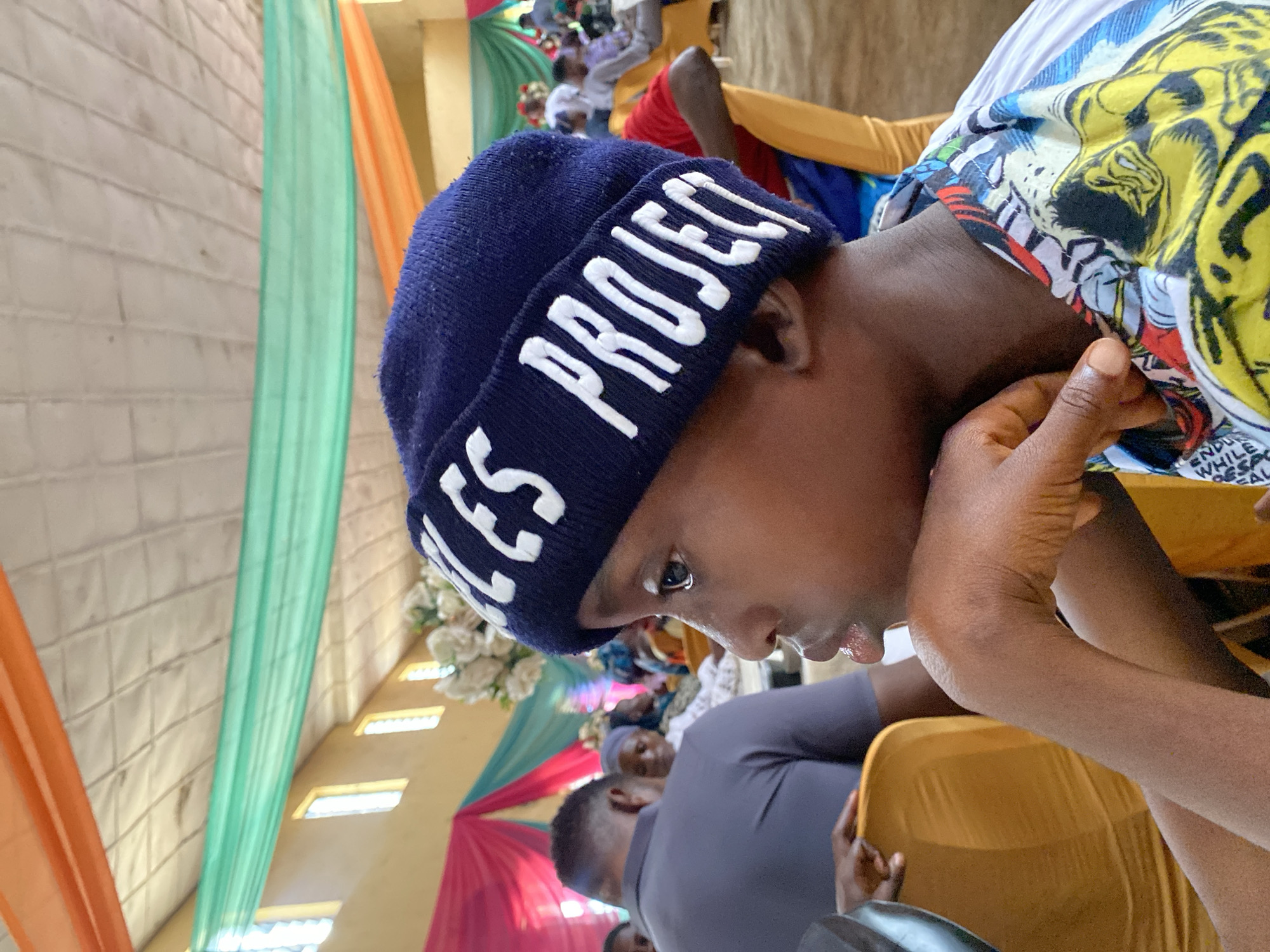 A medium shot captures a young person wearing a blue beanie with white lettering that reads "LES PROJECT," resting their chin on their hand and looking off to the side. The beanie's fabric has a textured knit pattern. The person's face is in profile, with a portion of their ear and neck visible. They are wearing a colorful shirt with a comic book-like print featuring yellow, blue, and red elements. The background suggests an indoor gathering, possibly a church hall or community center in Turu, Nigeria. The ceiling is adorned with orange and green draped fabric, and yellow windows are visible on the wall. Several other individuals are seated in the background, with some partially obscured by chairs. The overall lighting is ambient, with a slight overhead light source. The scene is framed from a slightly low angle, emphasizing the young person as the central subject.