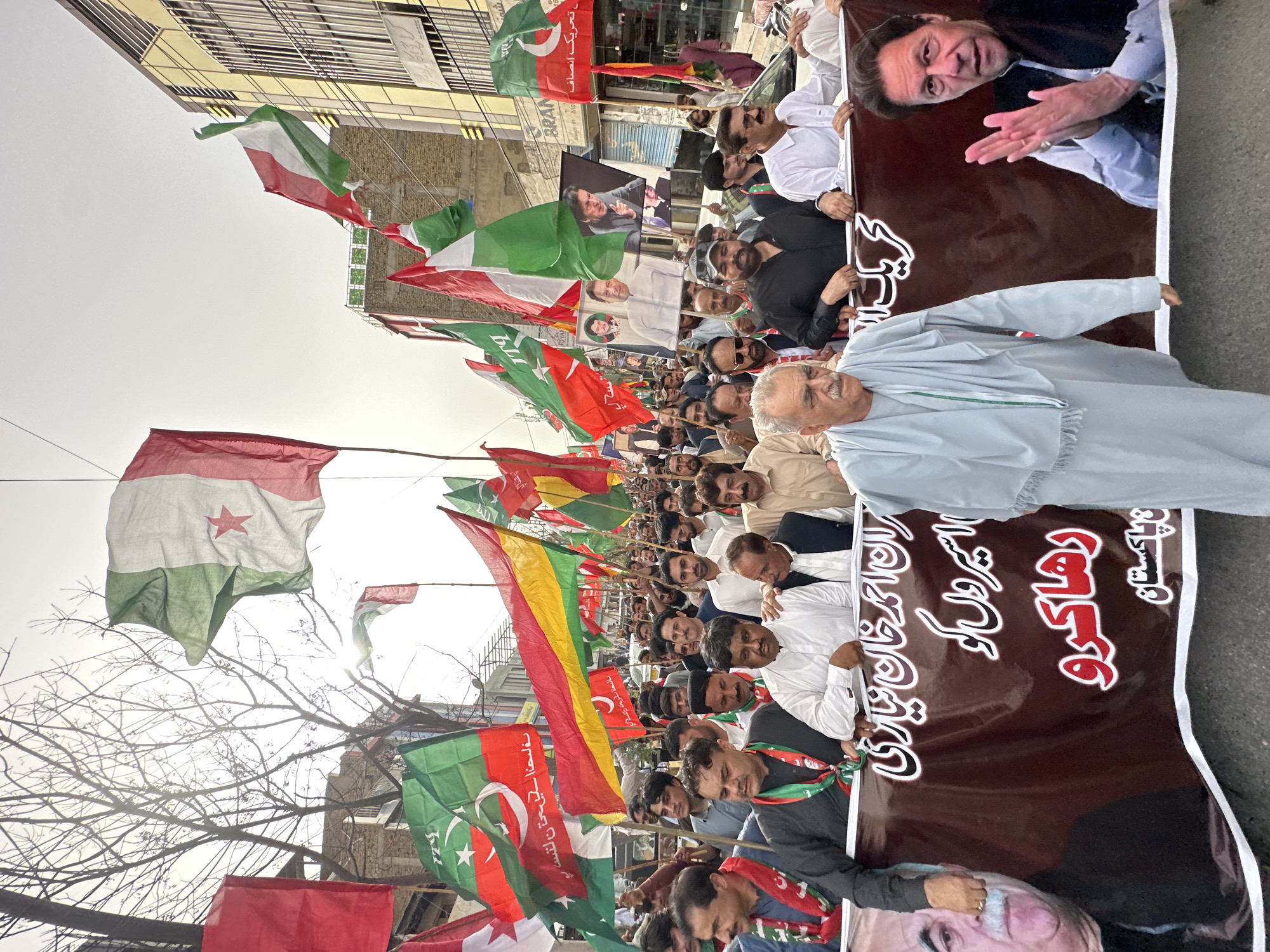 This is a ground-level, eye-level, medium shot photograph taken during daylight in Quetta City Tehsil, Pakistan. The scene depicts a large group of men participating in a street procession, likely a political rally, given the presence of numerous flags and banners. Many of the men are holding the flags aloft, which are predominantly green and white, with some featuring a star and crescent symbol, characteristic of the Pakistani flag, and others displaying a red star on a green and white background, possibly representing a specific political party.
A prominent banner is stretched across the foreground, held by several individuals. This banner features images of political figures, including what appears to be Imran Khan, and is adorned with text in Urdu script. The people in the procession are of various ages, and many are wearing traditional Pakistani attire, such as shalwar kameez. Some are wearing scarves or sashes, often in green and white. The expressions on their faces range from serious to engaged, suggesting active participation in the event.
The background of the photograph shows a city street lined with buildings of varying architectural styles. Some buildings have multiple stories and balconies, and there are visible power lines and streetlights. The sky is overcast, suggesting a cloudy or hazy day. The overall atmosphere appears to be one of organized public assembly. The procession is moving along the street, with people filling the width of the road.