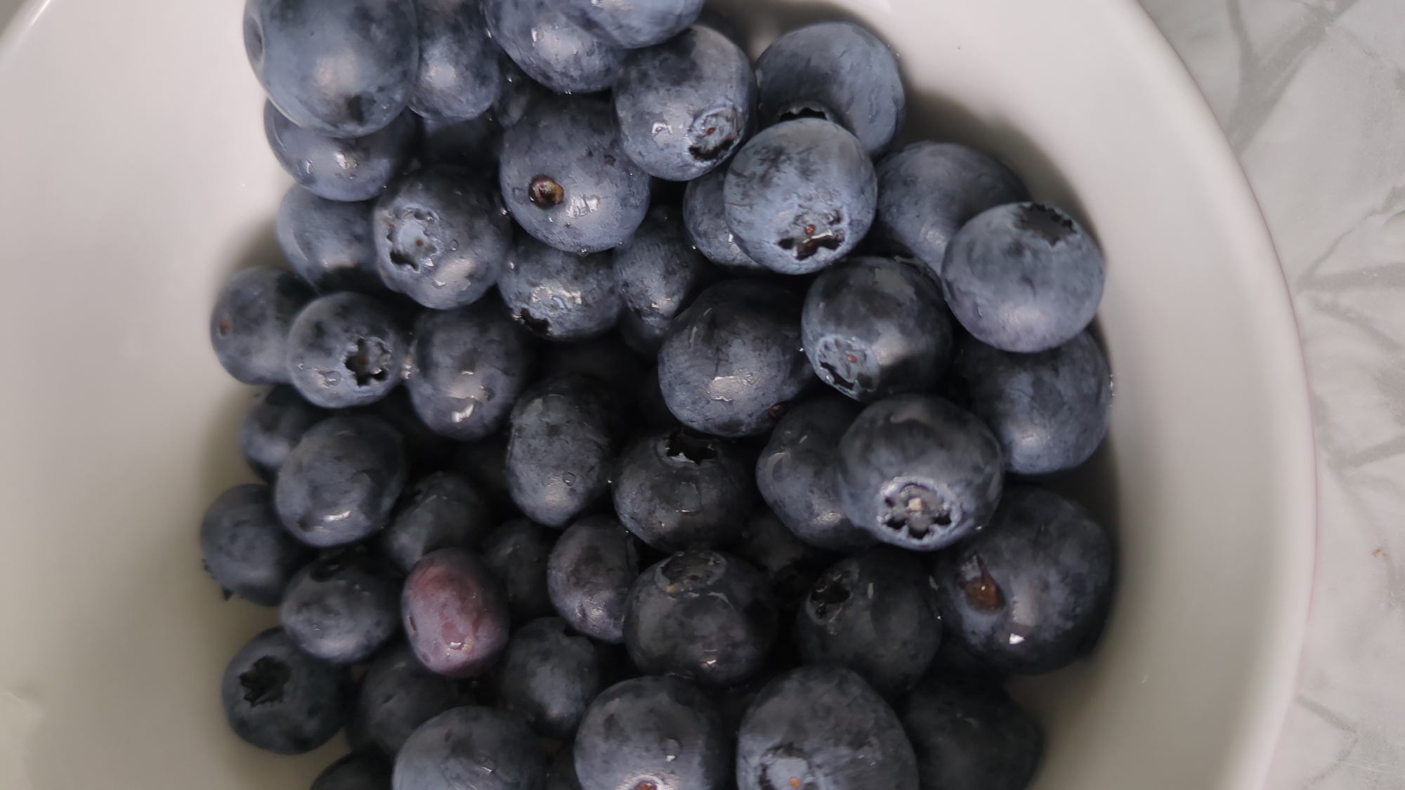 A close-up, overhead shot shows a white bowl filled with fresh blueberries. The blueberries are plump and round, with a deep blue-purple hue. Some of the blueberries have small water droplets on their surface, suggesting they have been recently washed. The bowl is white and smooth, and a hint of a light-colored, textured surface is visible beneath it. There are no people or any other objects present in the frame. The lighting is soft and diffused, highlighting the natural sheen of the fruit. The scene depicts a simple, fresh collection of blueberries.