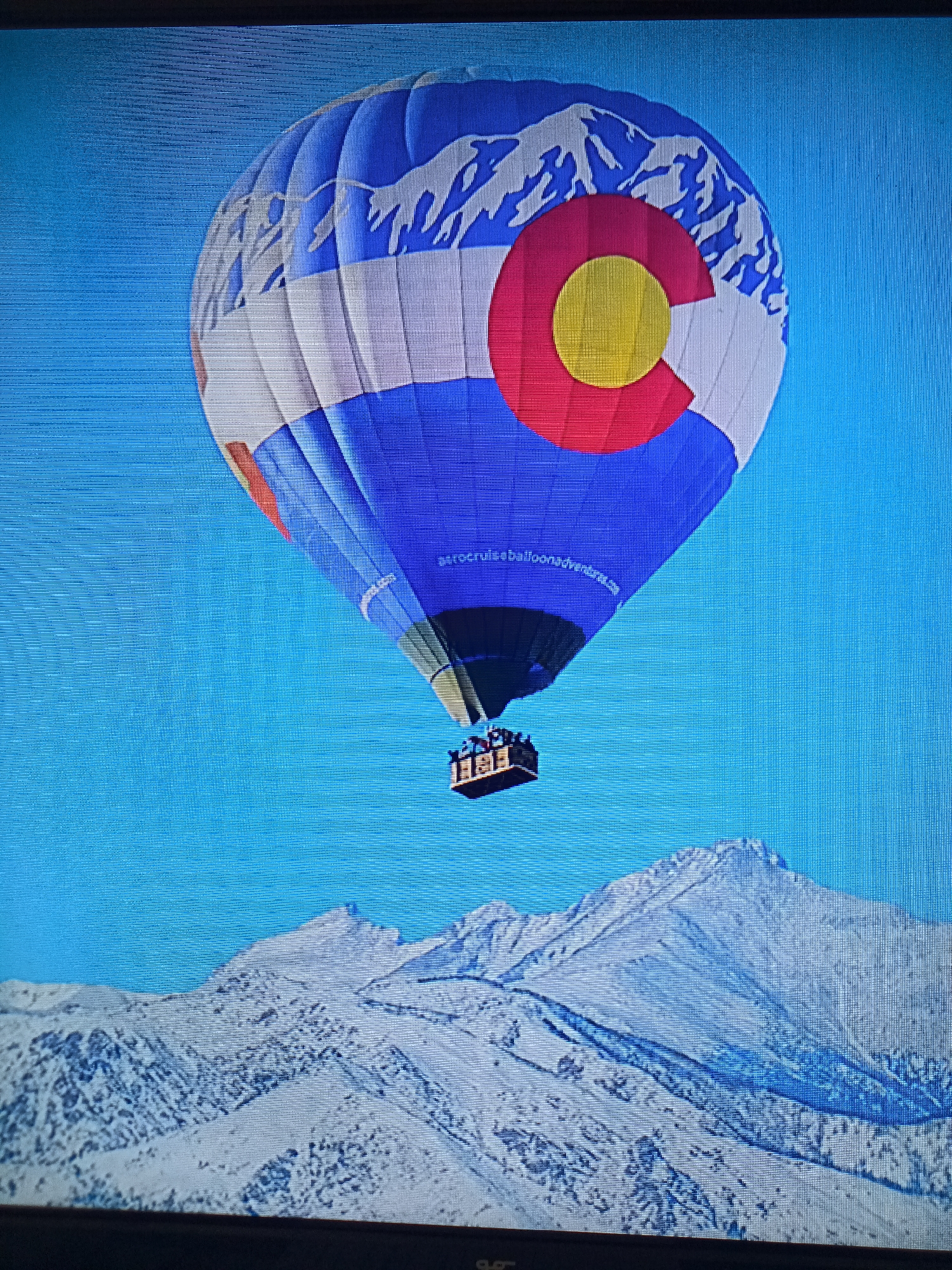 A hot air balloon, decorated with the Colorado flag design featuring white mountain peaks, a red circle, and a yellow circle against a blue background, floats in a clear blue sky. Below the balloon, a snow-covered mountain range is visible. The basket of the hot air balloon carries several passengers. A website address, "astrocruiseballoonadventures.com," is printed on the balloon. The scene appears to be a digitally altered image or a photograph of a screen displaying such an image, given the visible pixelation and screen artifacts. There is no indication of this image being located in Litomyšl, Czechia.