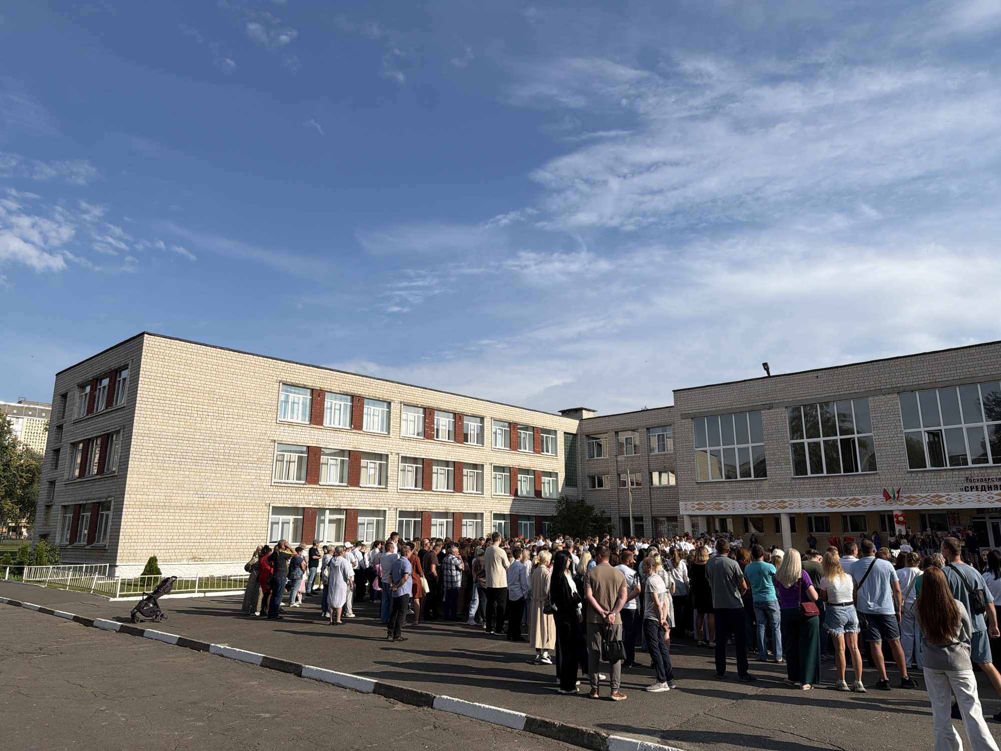 This is a full exterior, eye-level, wide shot of a large gathering of people in front of a two-story brick school building under a bright blue sky with scattered white clouds. The building has a light beige brick facade with rows of windows featuring red-trimmed frames on the left section and large, dark-paned windows on the right section. A white fence runs along the front of the building, bordering a paved area where a black stroller is parked. The crowd, composed of men, women, and children of various ages, is densely packed and facing the school building. Many are dressed in casual attire. The overall scene suggests an outdoor event or assembly taking place at the school.
