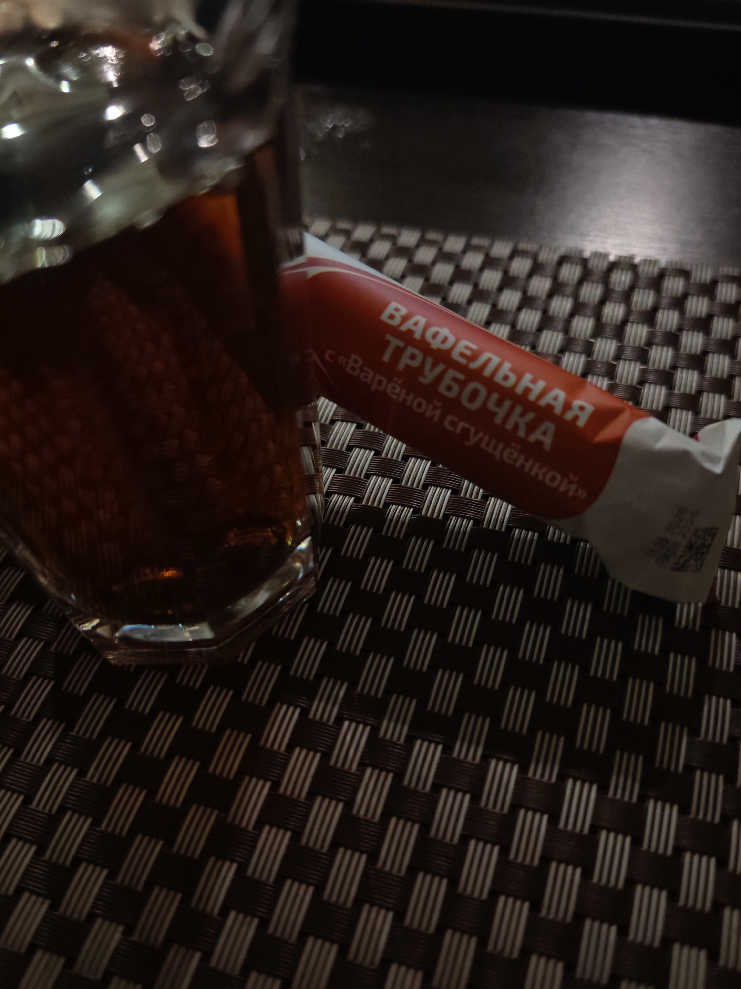 An overhead shot, in a close-up, features a clear glass with brown liquid and a foil wrapper. The wrapper is orange with white text, the liquid is dark amber, and the glass has a hexagonal base. Both the glass and wrapper are on a dark, woven placemat. There is no indication of the city or country.