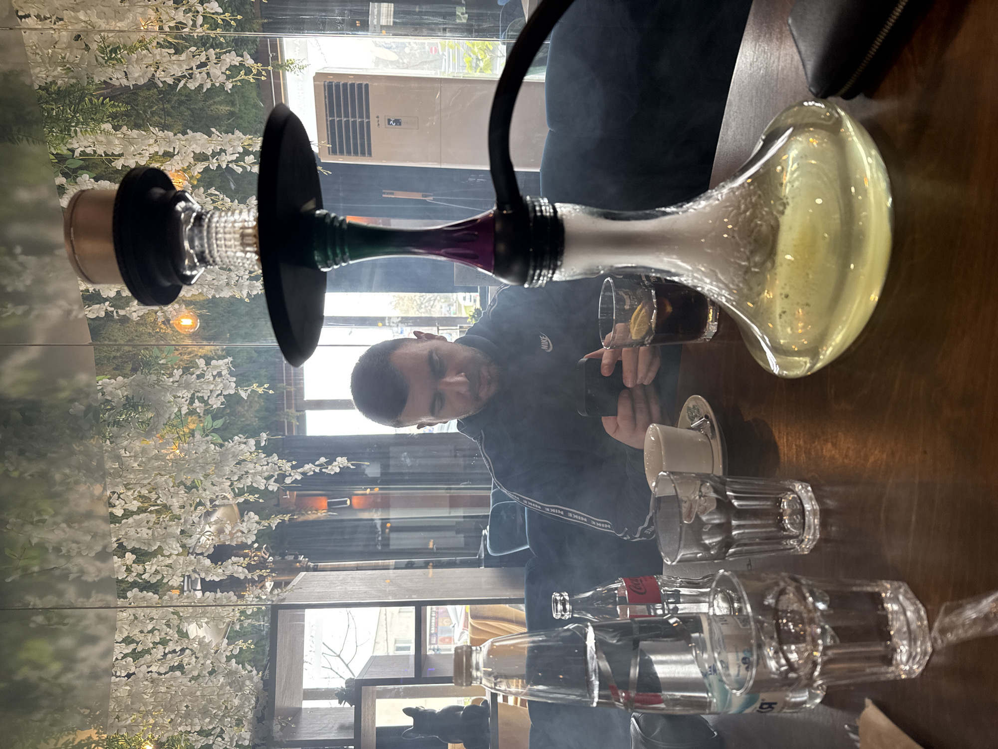 A man is seated at a table in an indoor setting that appears to be a restaurant or lounge, likely in Sofia, Bulgaria. The man has short dark hair and is wearing a dark Nike tracksuit. He's holding a dark liquid in a glass, possibly a mixed drink, and appears to be smoking a hookah that is set up on the table. There is smoke visible in the scene. The hookah has a bulbous glass base filled with a yellow liquid. Other objects on the table include a small coffee cup and saucer, two empty glasses, and two bottles. Behind the man is a mirrored wall with decorative white floral arrangements. Outside the window, a street scene is visible, suggesting an urban setting. There is an air conditioner visible outside the window. The table surface appears to be wooden or a wood-like material. The overall lighting of the scene is subdued, creating a relaxed atmosphere.