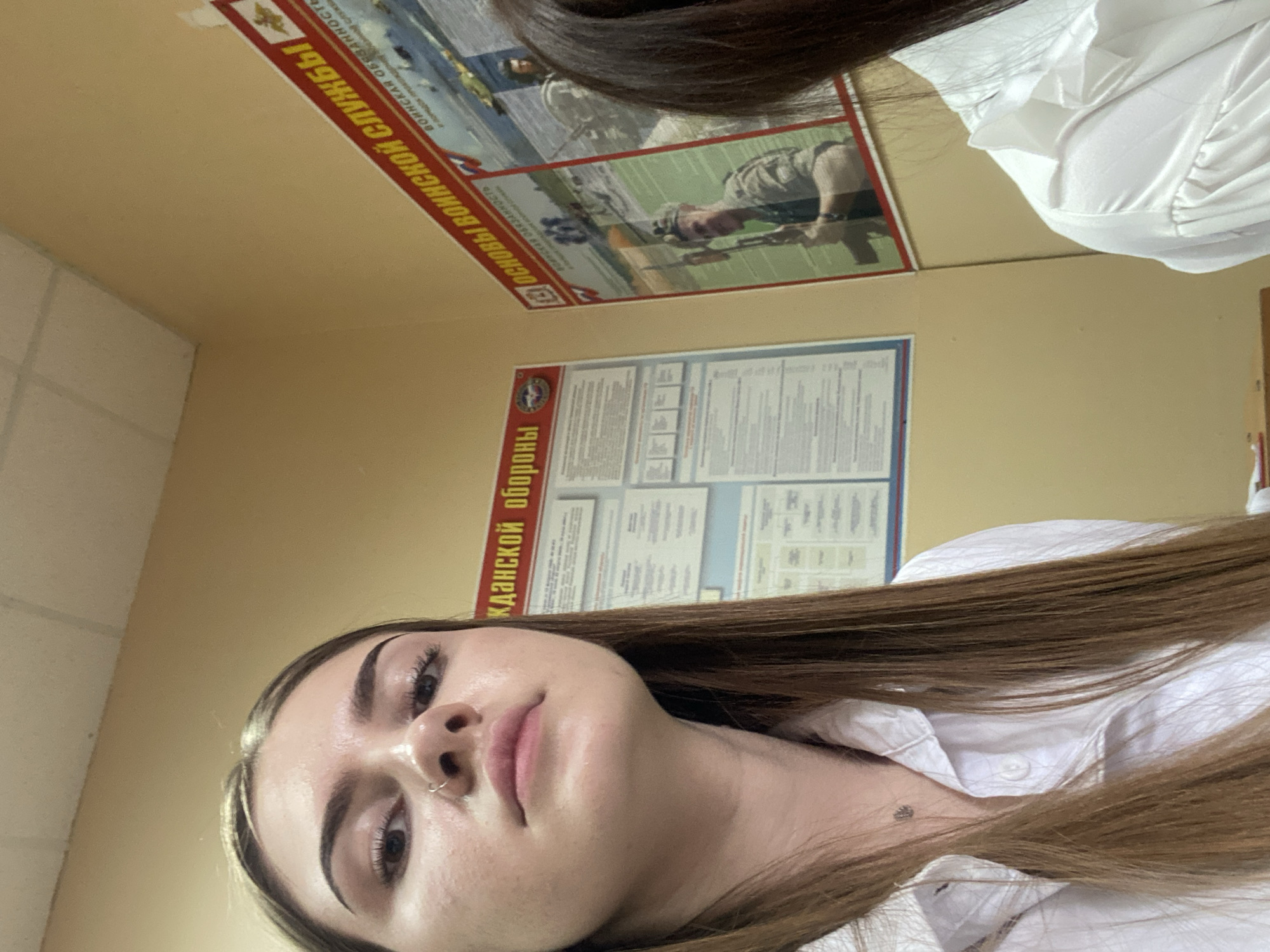 A medium shot captures a young woman with long, brown hair, wearing a white button-up shirt. She is positioned in the foreground, with her face angled towards the camera. Her expression is neutral, and she has a nose ring. Behind her, on the wall, are two posters. The poster on the left has red text that reads "ГРАЖДАНСКОЙ ОБОРОНЫ" (Civil Defense). The poster on the right, also with red text, says "ОСНОВЫ ВОИНСКОЙ СЛУЖБЫ" (Fundamentals of Military Service) and features images of military personnel. The wall appears to be a light yellow color, and the ceiling is white with visible ceiling tiles. The environment suggests an indoor setting, potentially a classroom or office space within a building in Dyakovo, Russia. Another person's dark hair is visible on the far right side of the frame, partially obscuring the rightmost poster.