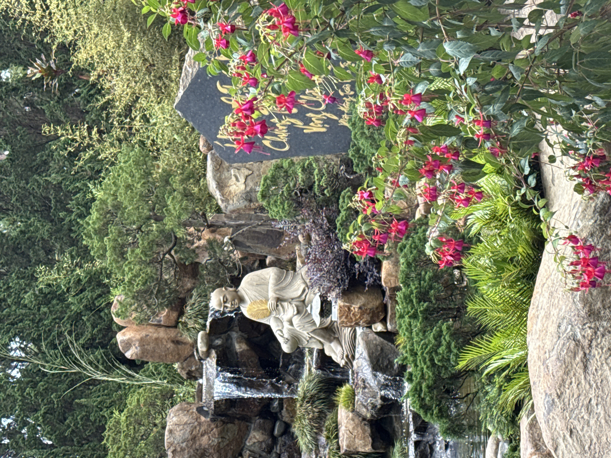 The image captures a garden scene in Da Lat, Vietnam. A stone statue of a seated figure, possibly a monk, is positioned within a small waterfall feature constructed of rocks. To the right, vibrant fuchsia flowers cascade near a large boulder. A plaque with golden lettering, partly obscured by the flowers, sits atop a rock formation. Dense greenery surrounds the scene, and the perspective is slightly tilted.