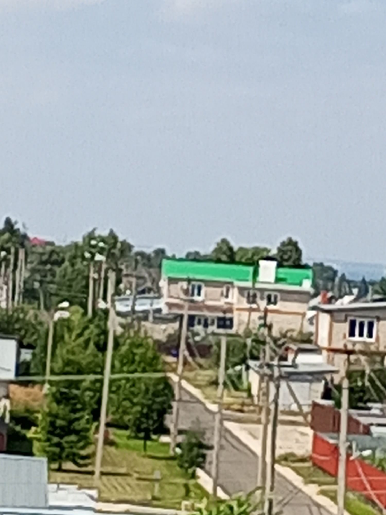 The image shows a residential area with several buildings and trees under a clear sky. In the foreground, there are utility poles with wires. To the right, a two-story building with a green roof and white trim is visible. Adjacent to it, another building with a light-colored facade has windows and a red roofline in the background. Further to the right, a smaller building with a white facade and dark rectangular windows is present. The area appears to be a suburban or rural setting.